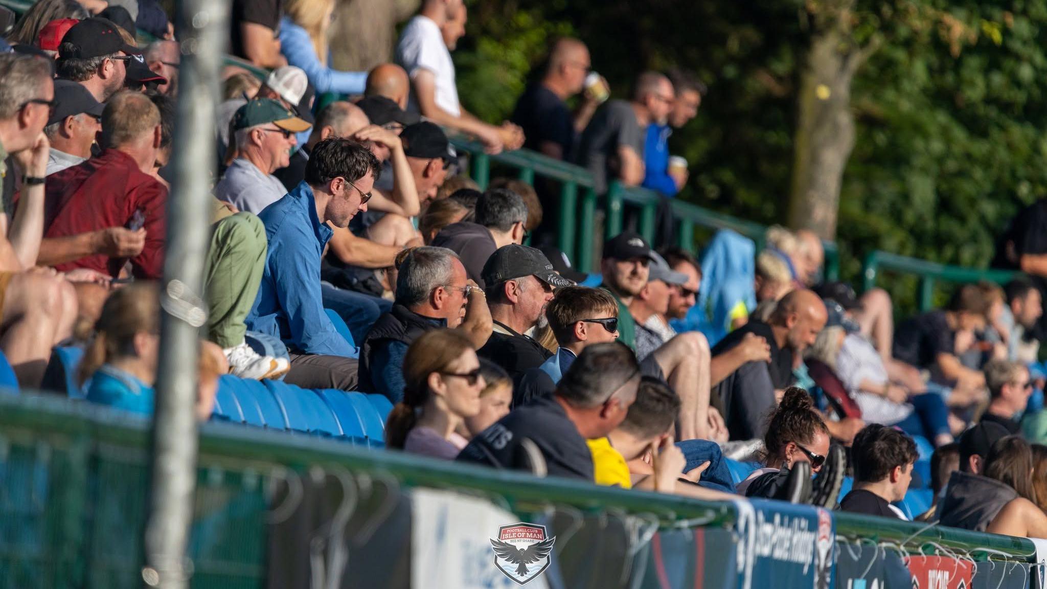 Men and women sit in the stands of a football ground, wearing sunglasses and hats, and look towards the pitch.