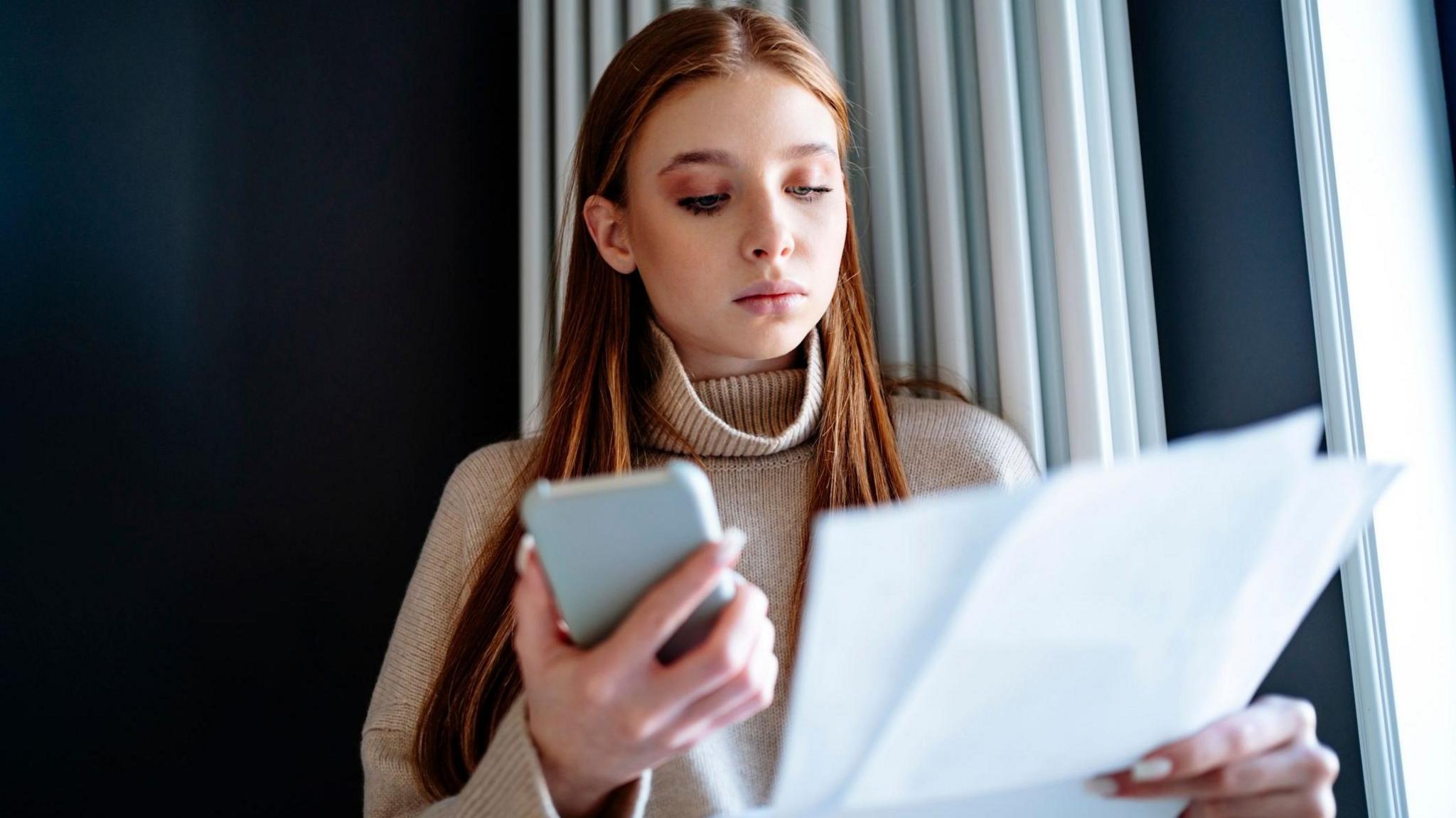 Young woman stands in front of a radiator holding a smartphone and a paper bill.