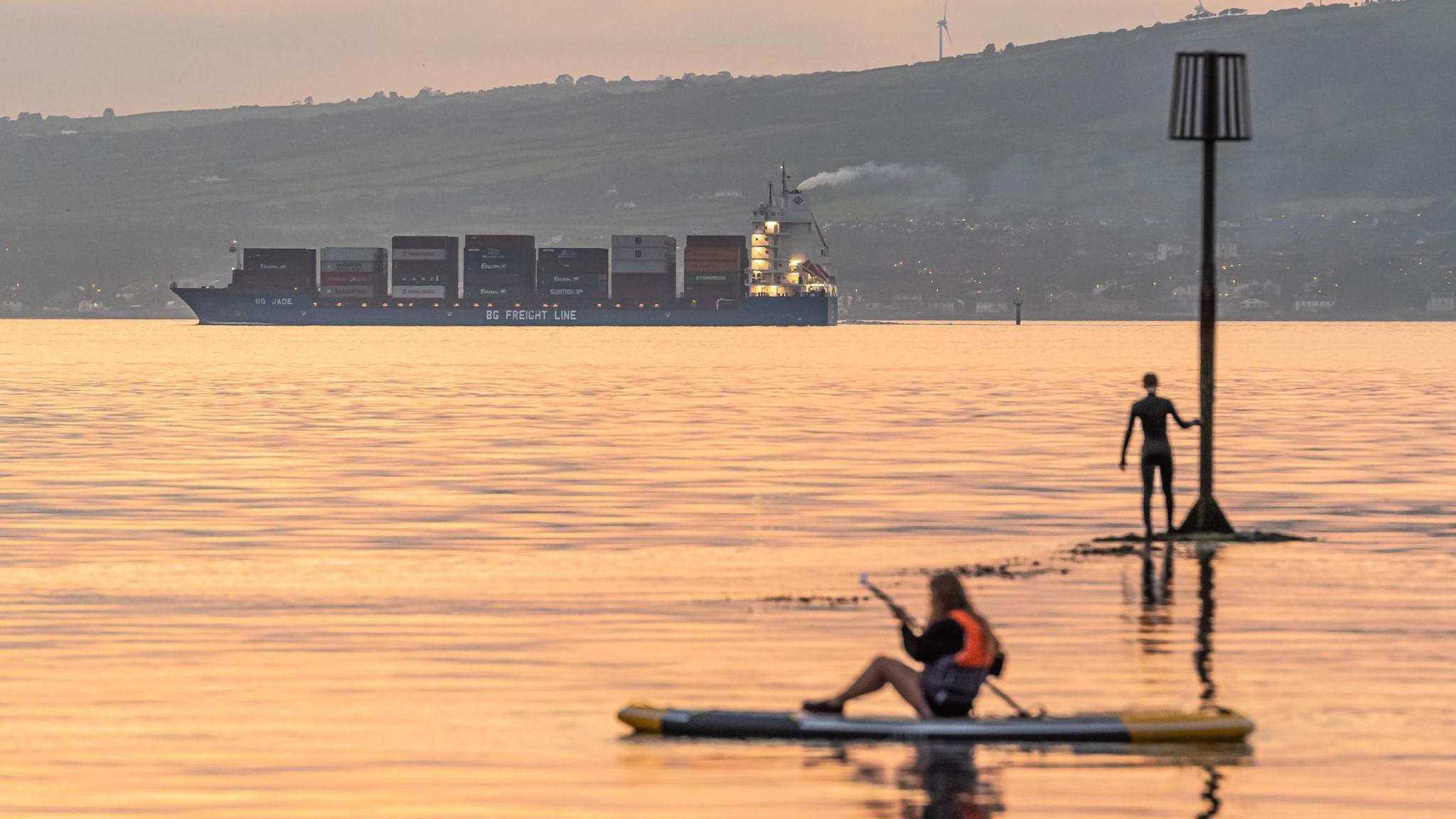 A cargo ship is sailing from right to left of the frame as two paddle-boarders are viewed in the foreground. It's evening and the water has an orange reflection to it. A large green hill sits behind the ship.