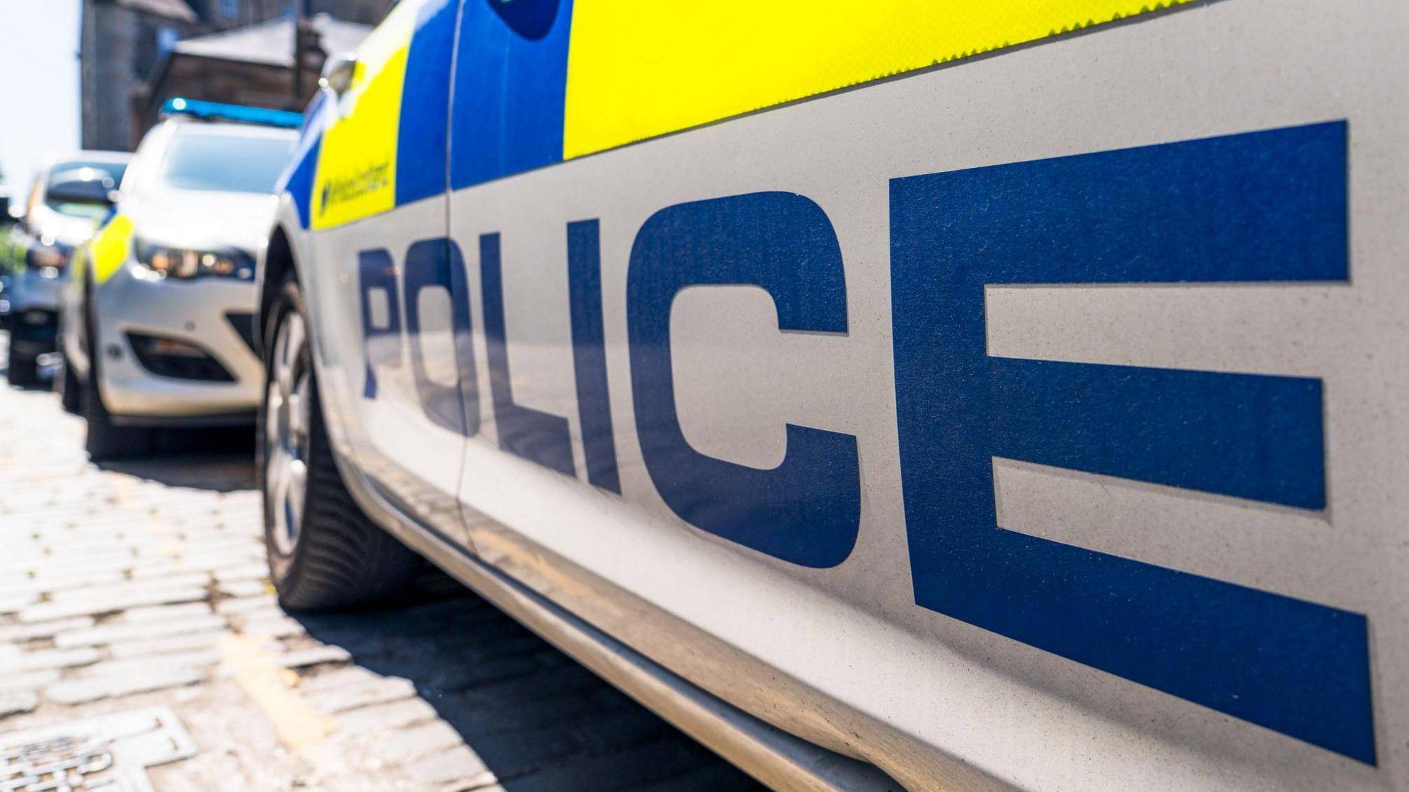 Generic image of the side of a police car. The word POLICE font is printed under fluorescent decals on a UK street, with other police vehicles in the background.