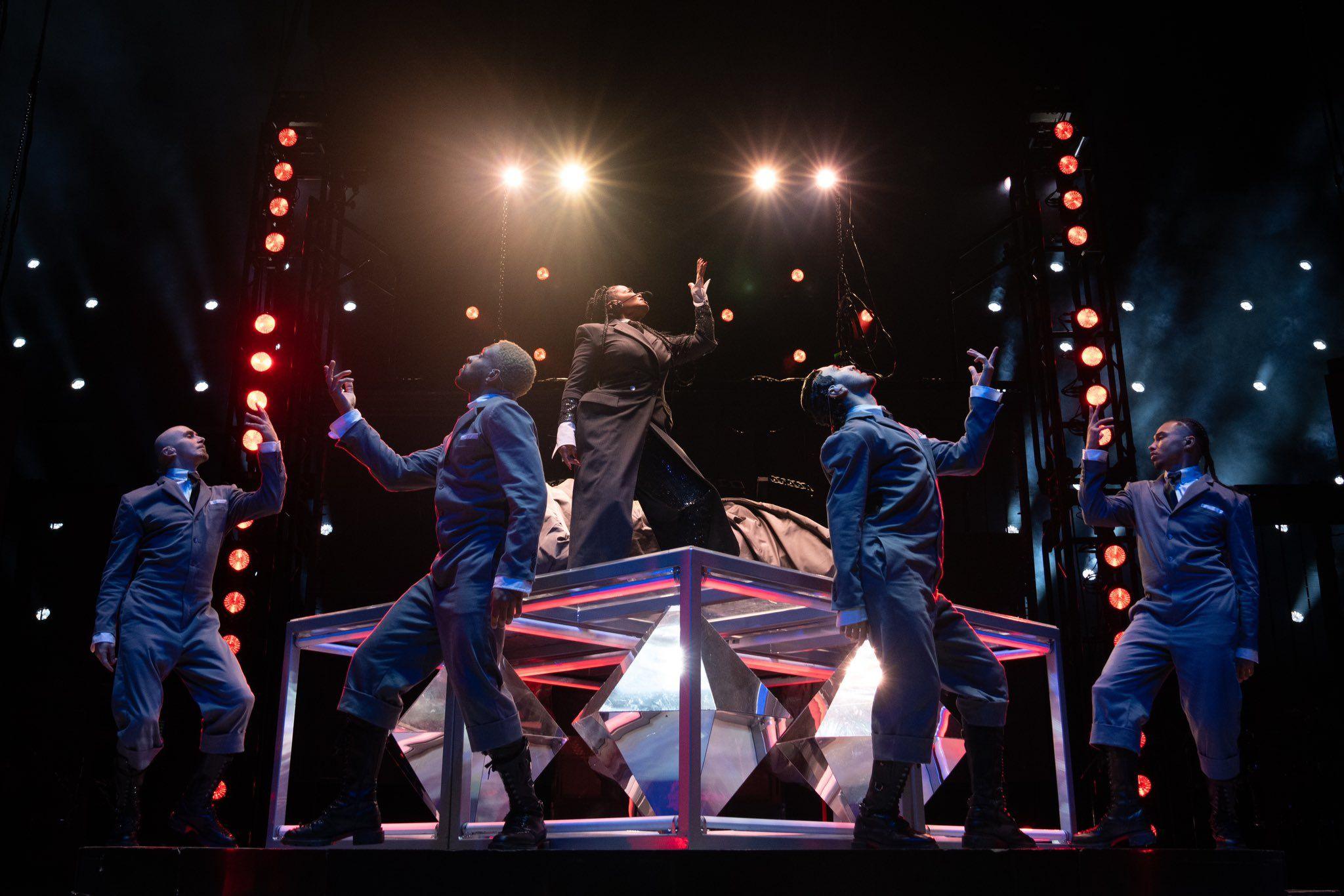 Janet Jackson on stage during the Together Again tour. She poses on a podium while four male dancers mirror her on the stage