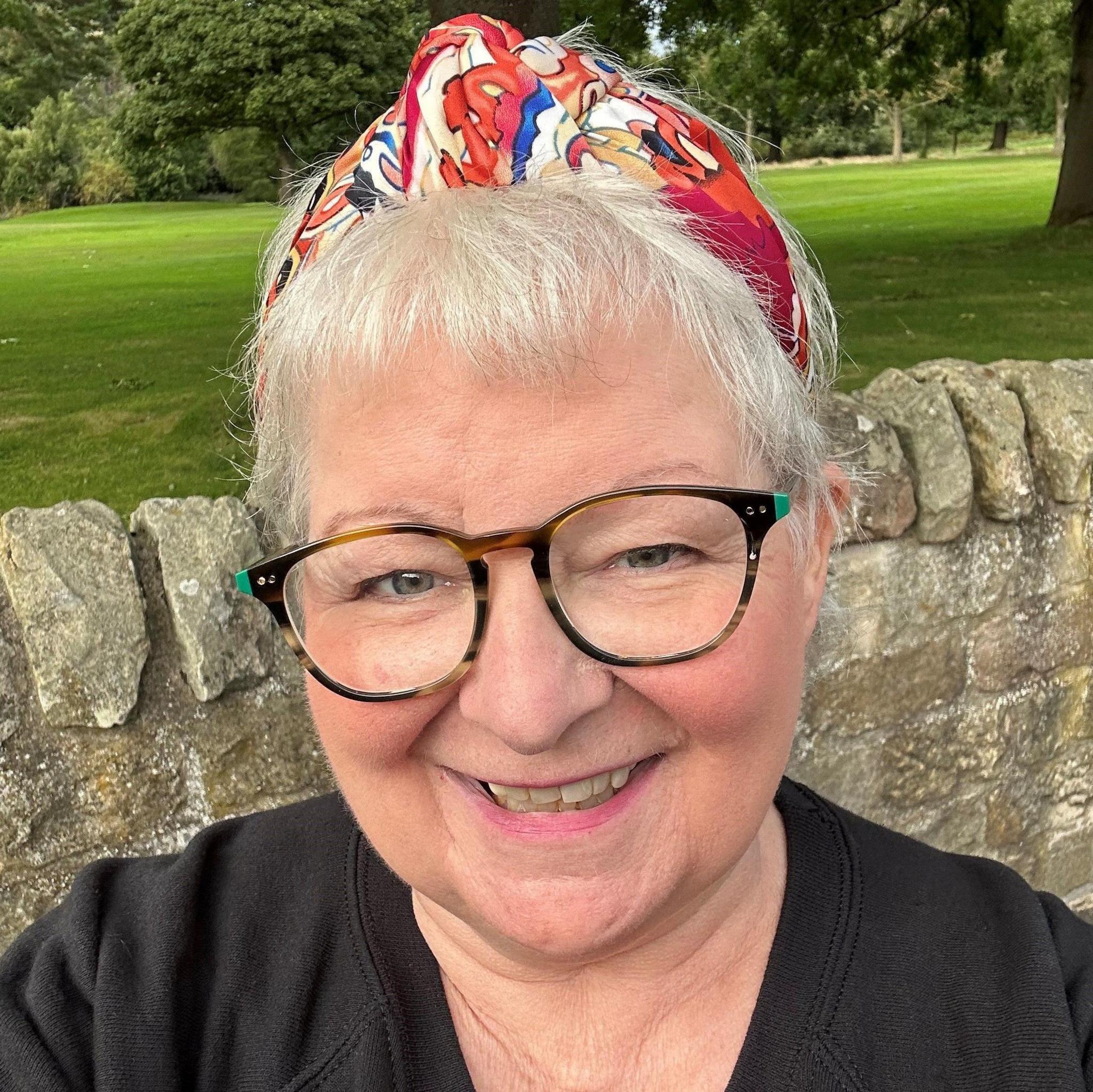 Janey Godley, with short grey hair, dark glasses and a red bandana on her head, smiles at the camera with a stone wall and green park behind her 