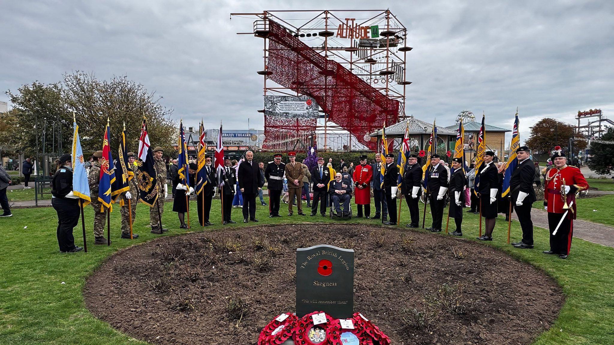 A circular piece of brown soil with a plaque in front of it with a red poppy and 'The Royal British Legion Skegness' printed on to it with wreaths in front of it. Standing around the circle are several cadets holding Union Jack flags. Behind them is a high-wire attraction with a red sea of poppies draped from it.