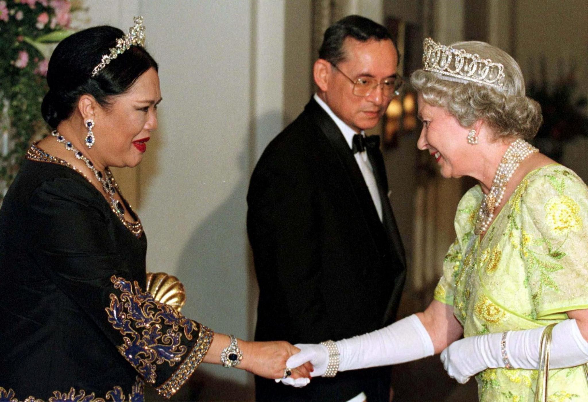 Queen Sirikit, wearing a black dress, greets Queen Elizabeth II, wearing a light freen dress and white gloves. Thailand's late King Bhumibol stands in the background between them. 