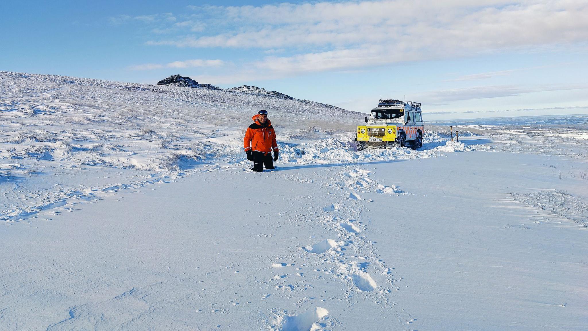 A rescuer stands with snow up to his thighs with the search and rescue team's Land Rover behind him. There are deep foot prints leading from the man to the person taking the photograph. There is blue sky and snow covers everywhere in sight. 