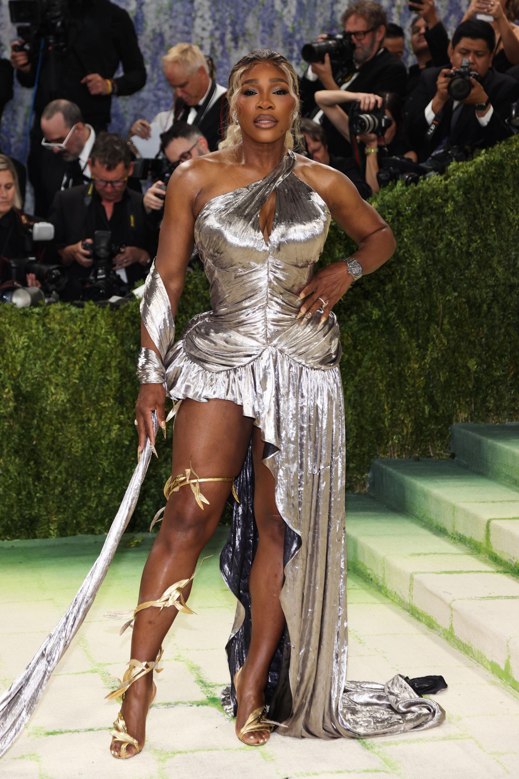 US retired professional tennis player Serena Williams poses on the red carpet wearing a silver shiny dress what has one leg covered by the skirt and the other showing. 