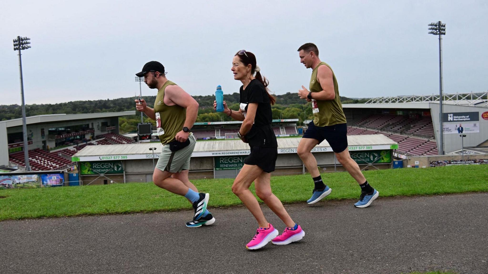 Three runners moving from right to left from the perspective of the camera are on a hill overlooking Northampton Town's Sixfields Stadium