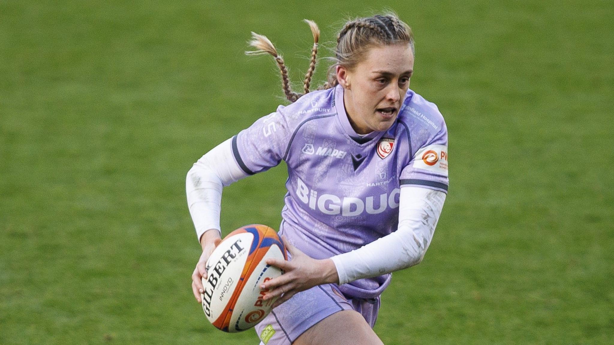 Gloucester-Hartpury centre Hannah Dallavalle wearing a purple Rugby shirt and carrying the ball during a Premiership Rugby game. 