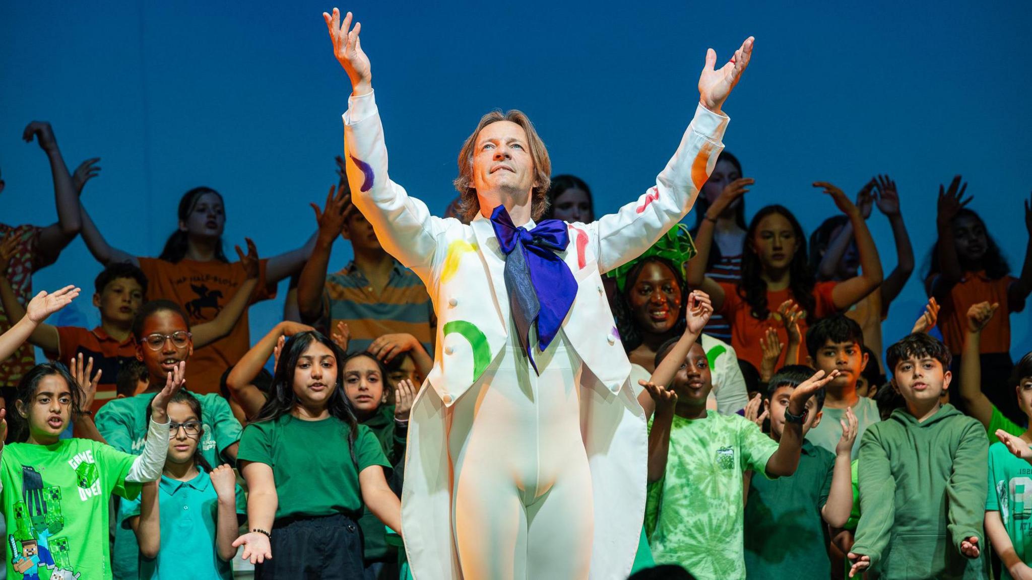 Male dancer dressed in white costume with green, yellow and orange pattern with blue cravat surrounded by children in green tops