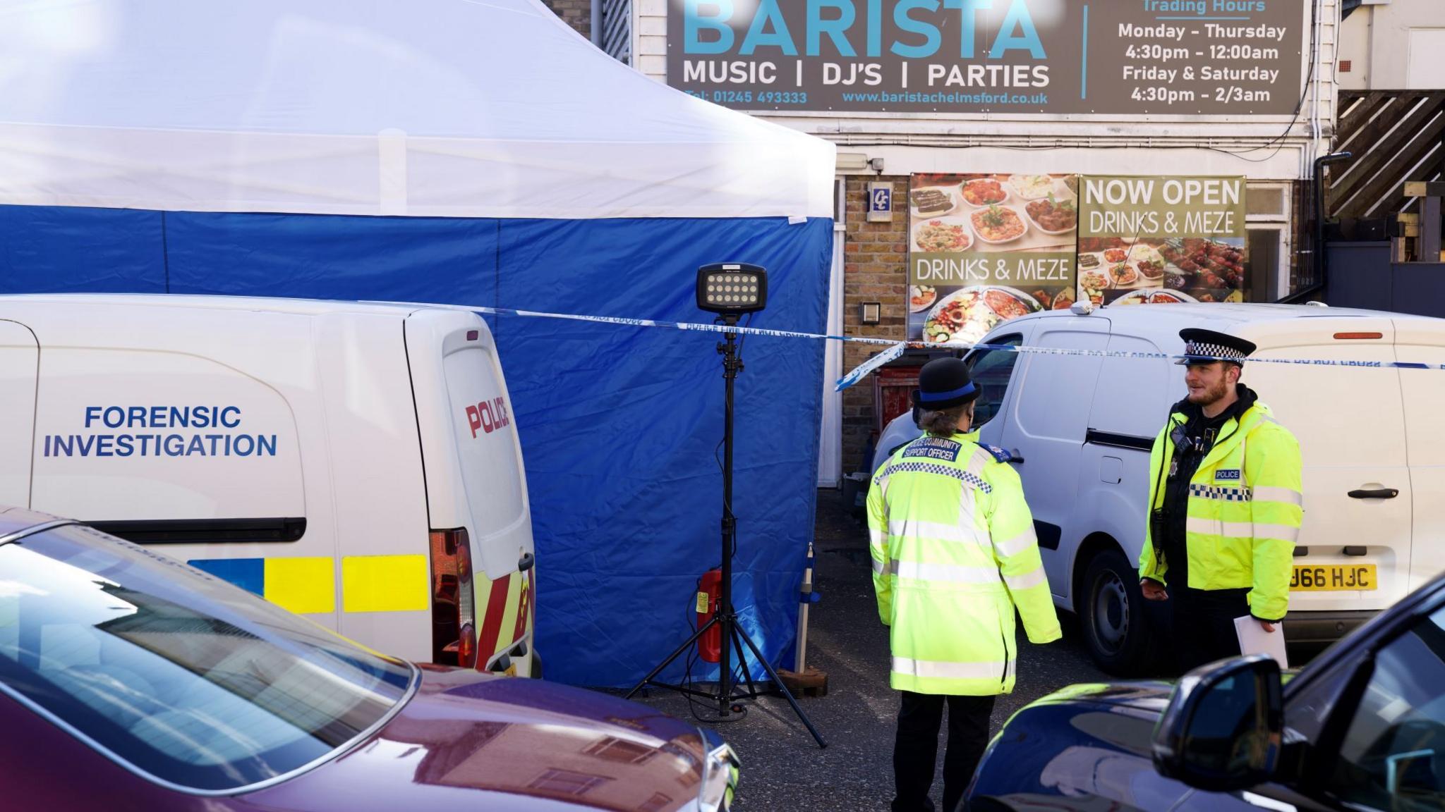 Police officers wearing hi-vis jackets guard the entrance to a shop in Chelmsford. Behind them is a large blue and white tent and police tape. There are several vans and cars parked in front of the building.