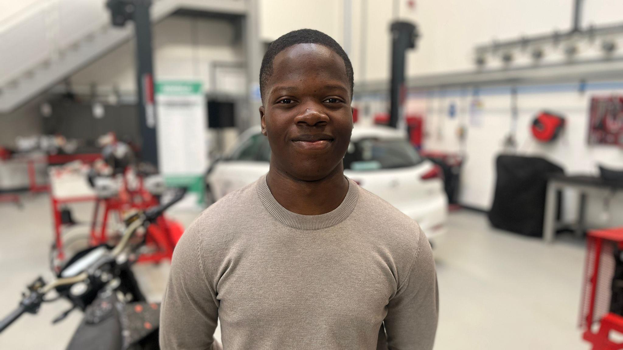 Simba is stood in a workshop at his college, smiling into the camera centre of frame. He is wearing a stone coloured jumper, and blurred in the background behind him is a car, a motorbike and several work benches.