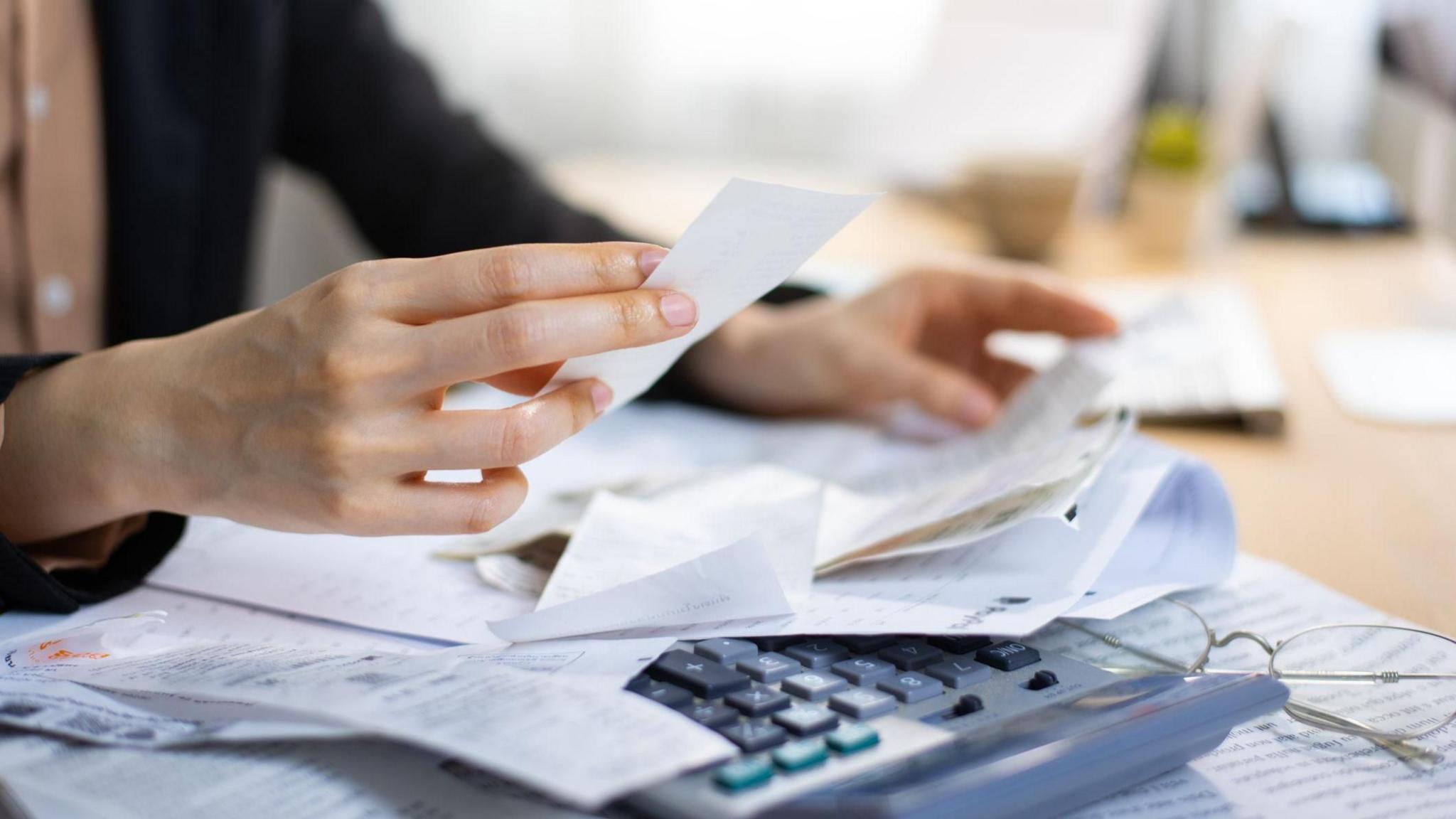 A close-up of someone looking at household bills with a calculator in front of them on a desk next to a pair of glasses