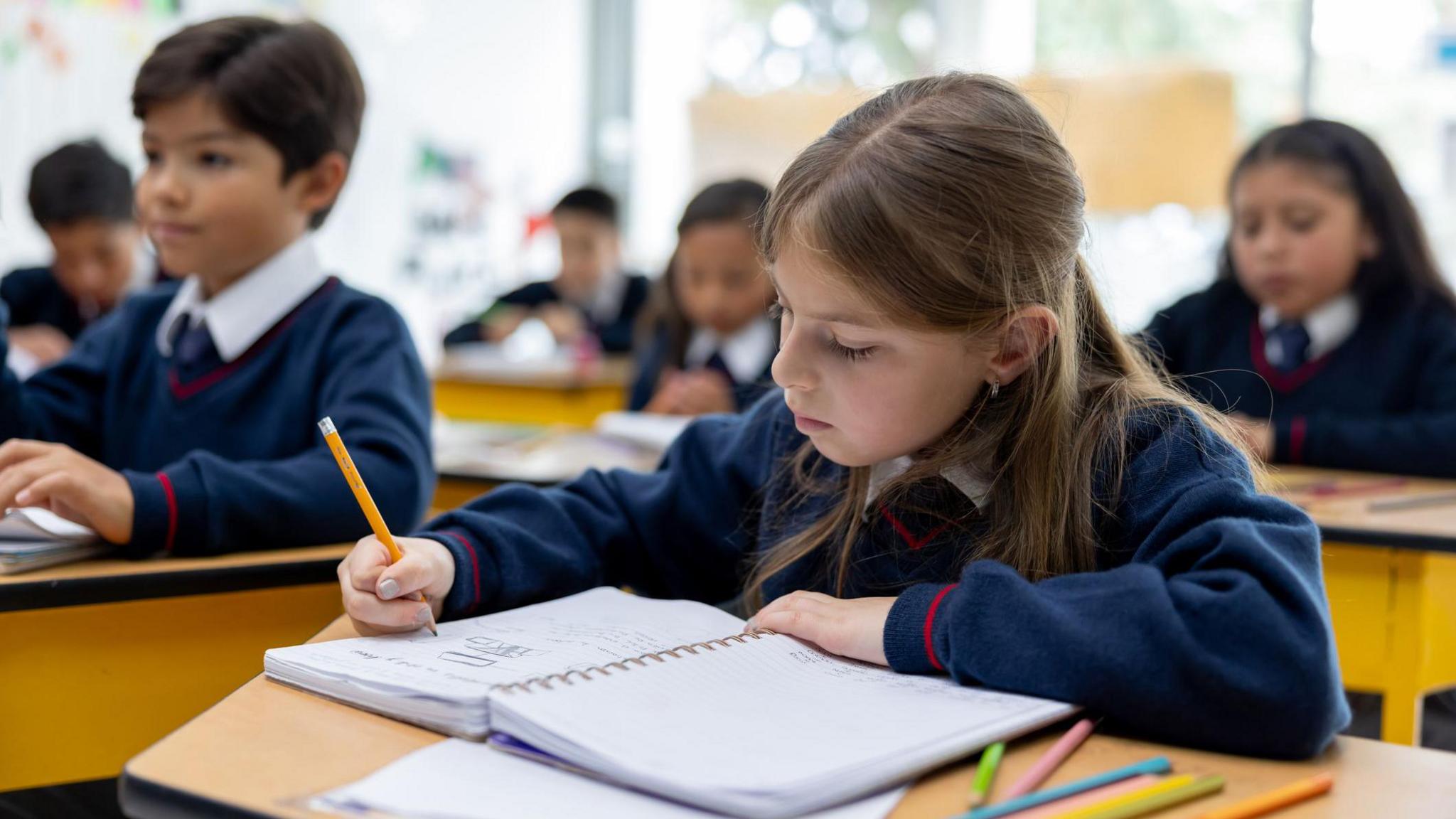 a girl in a navy school uniform writing in her school book at a wooden desk, with classmates out of focus in the background