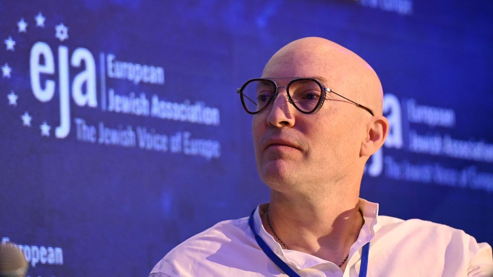 Alex Hearn wearing glasses, a white shirt and a lanyard, looks to the left of the camera lens. Behind him is an advert for the European Jewish Association.  