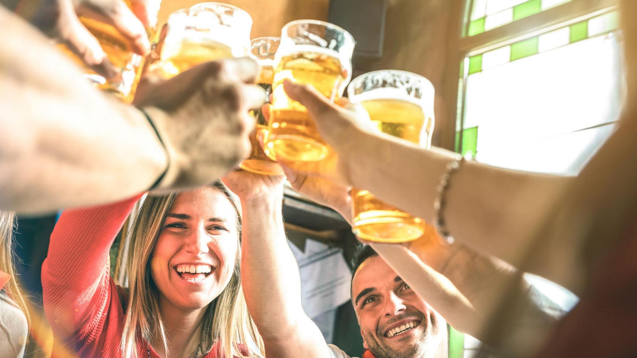 A young woman and a man smile as a large number of arms holding pint glasses of lager or beer are held above their heads, as if they are saying cheers