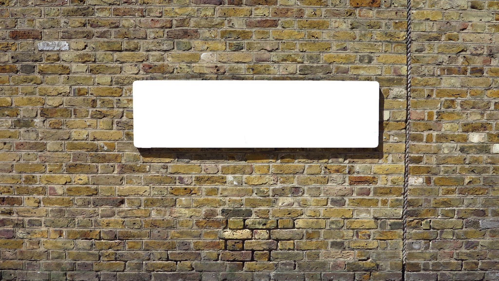 A blank road sign on a brick wall. The bricks are yellow and brown in colour. The road sign is a blank white slate where a name would usually be.