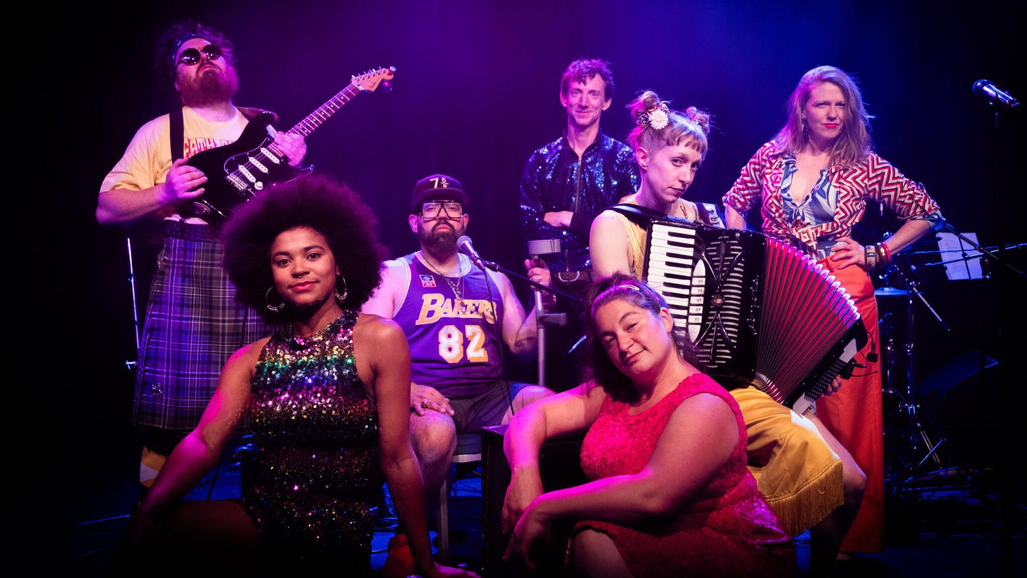 A group of performers from a theatre company. In the group of seven, there is a mixture of men and women. Some of them are holding musical instruments and all of them are looking at the camera and posing. They are standing under purple spotlights.