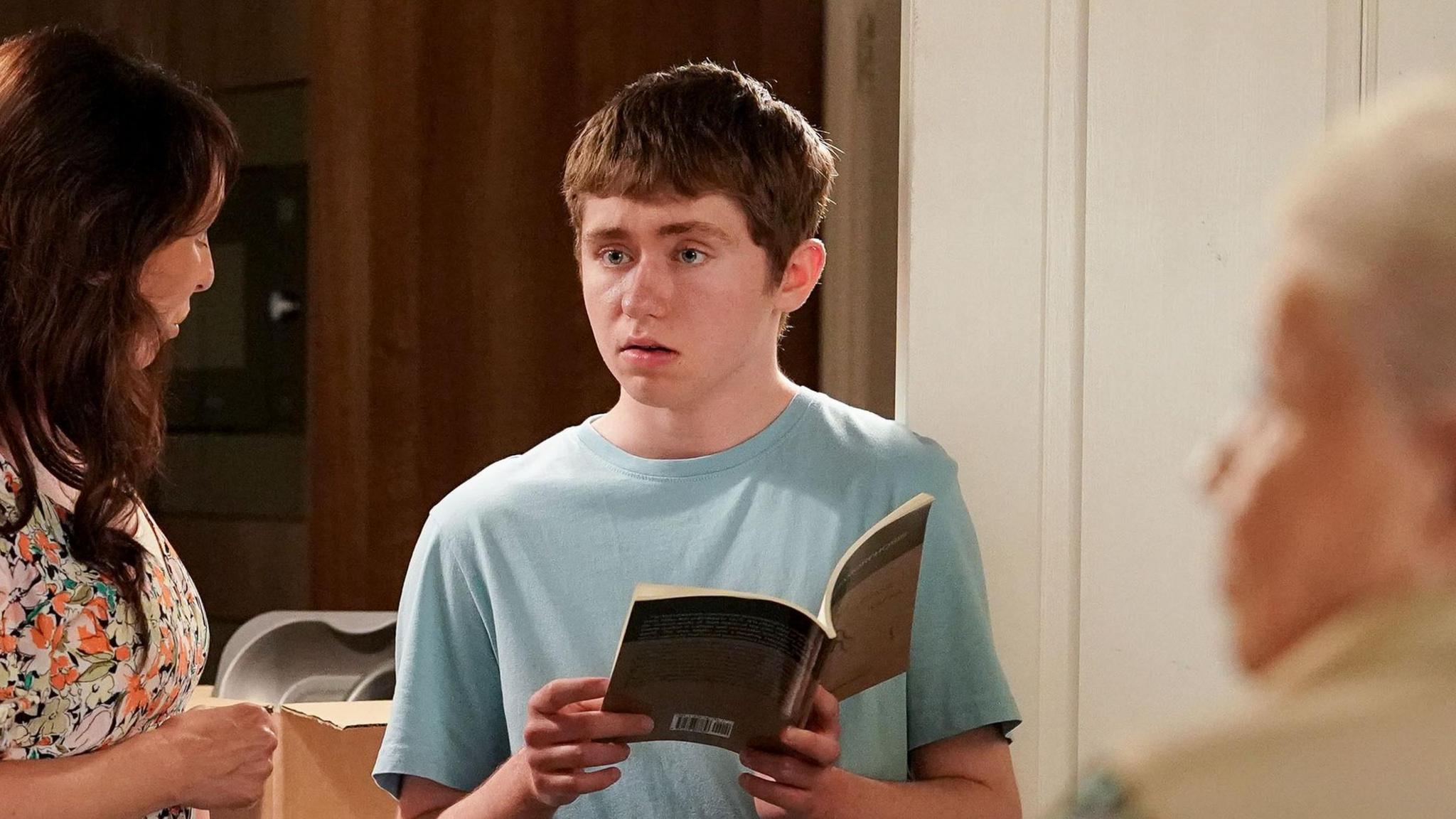 Barney Mitchell, played by Lewis Bridgeman, is stood holding a book and dressed in a blue t-shirt. He is stood next to Honey Mitchell and Big Mo.