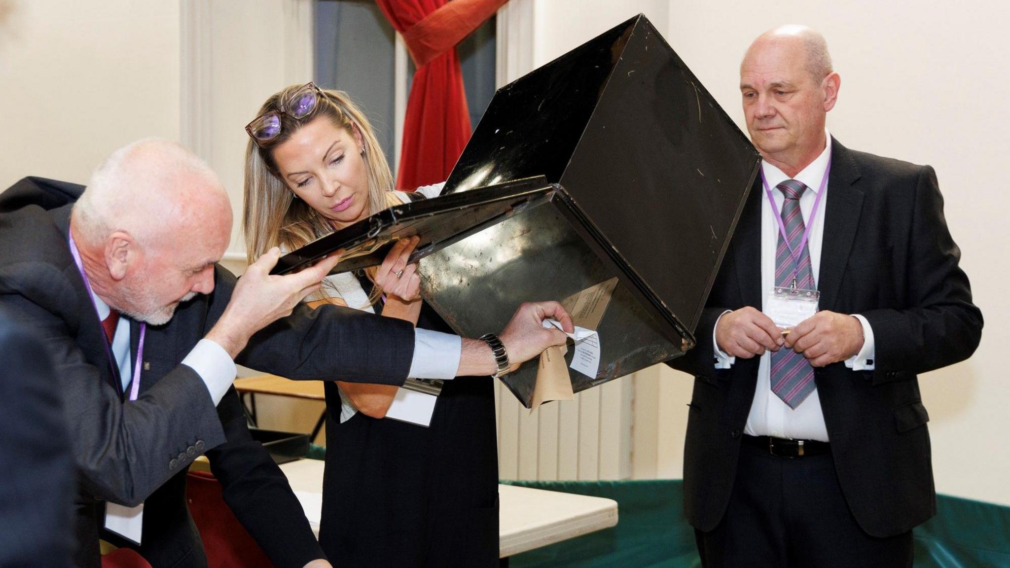 Three people pouring votes out of a ballot box. The two men are wearing dark suits and the woman, in the centre, is wearing a dark dress. 