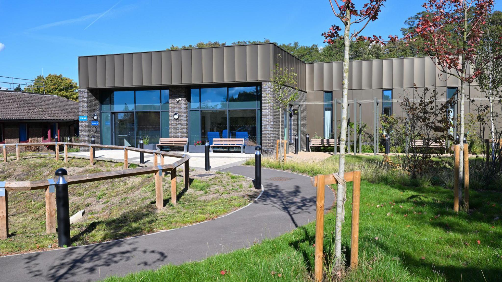 A single-storey building covered in brown panels with large glass doors on the left. In front of the building is a black path leading to the door, and a grassy area. Behind the building are trees and a blue sky.
