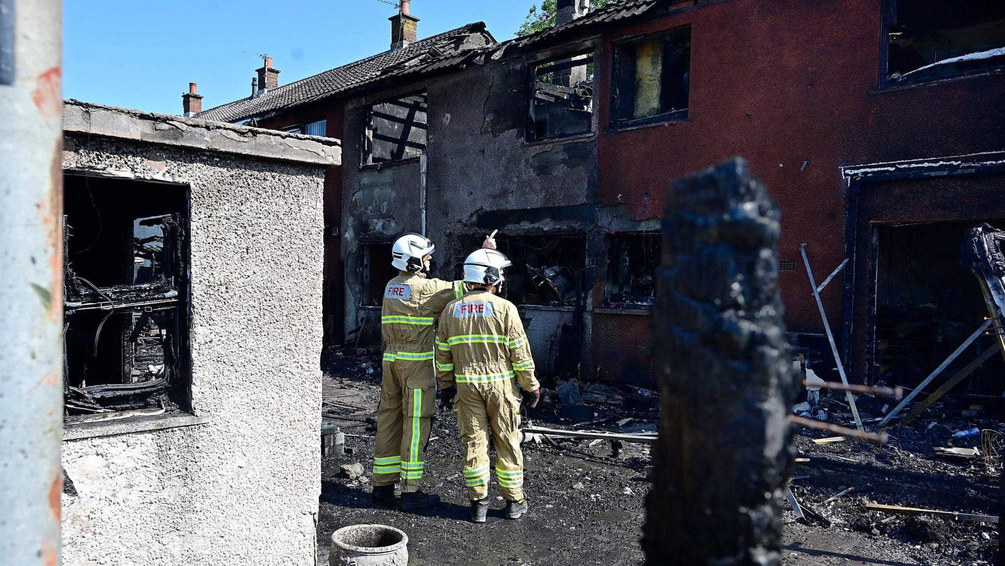 Two firefighters in yellow uniforms and white helmets in front of the burnt out red and white houses. 
