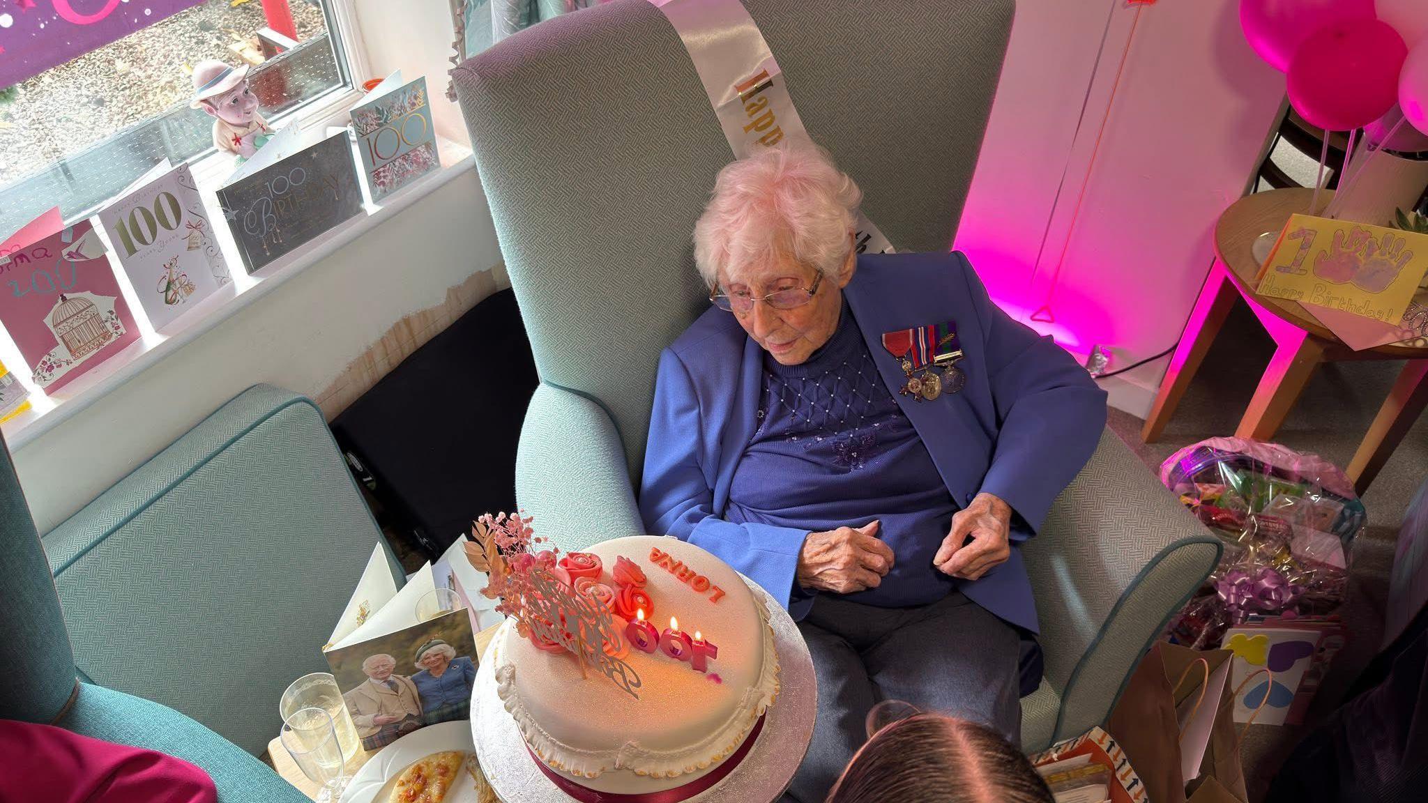 An elderly woman with white hair and glasses sits in an armchair, wearing a blue top and jacket with military medals, and surrounded by 100 birthday cards and balloons, including a card with a picture of King Charles and Queen Camilla. She has a white birthday cake in front of her with pink writing reading "Lorna" and pink candles and flowers.