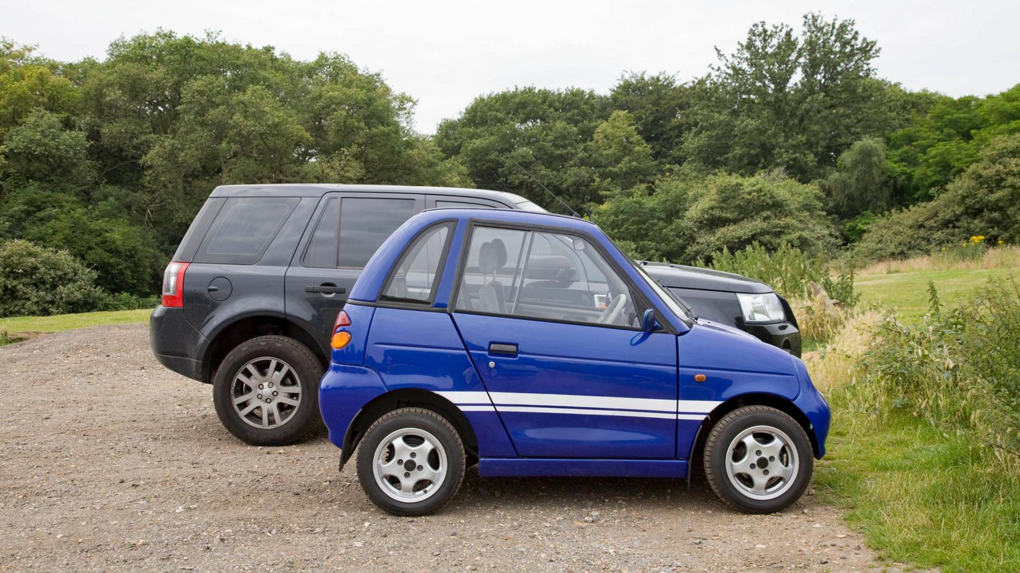 A small blue car parked in a park car park, in front of a larger grey SUV. The difference in length and height of the two cars is clearly visible.