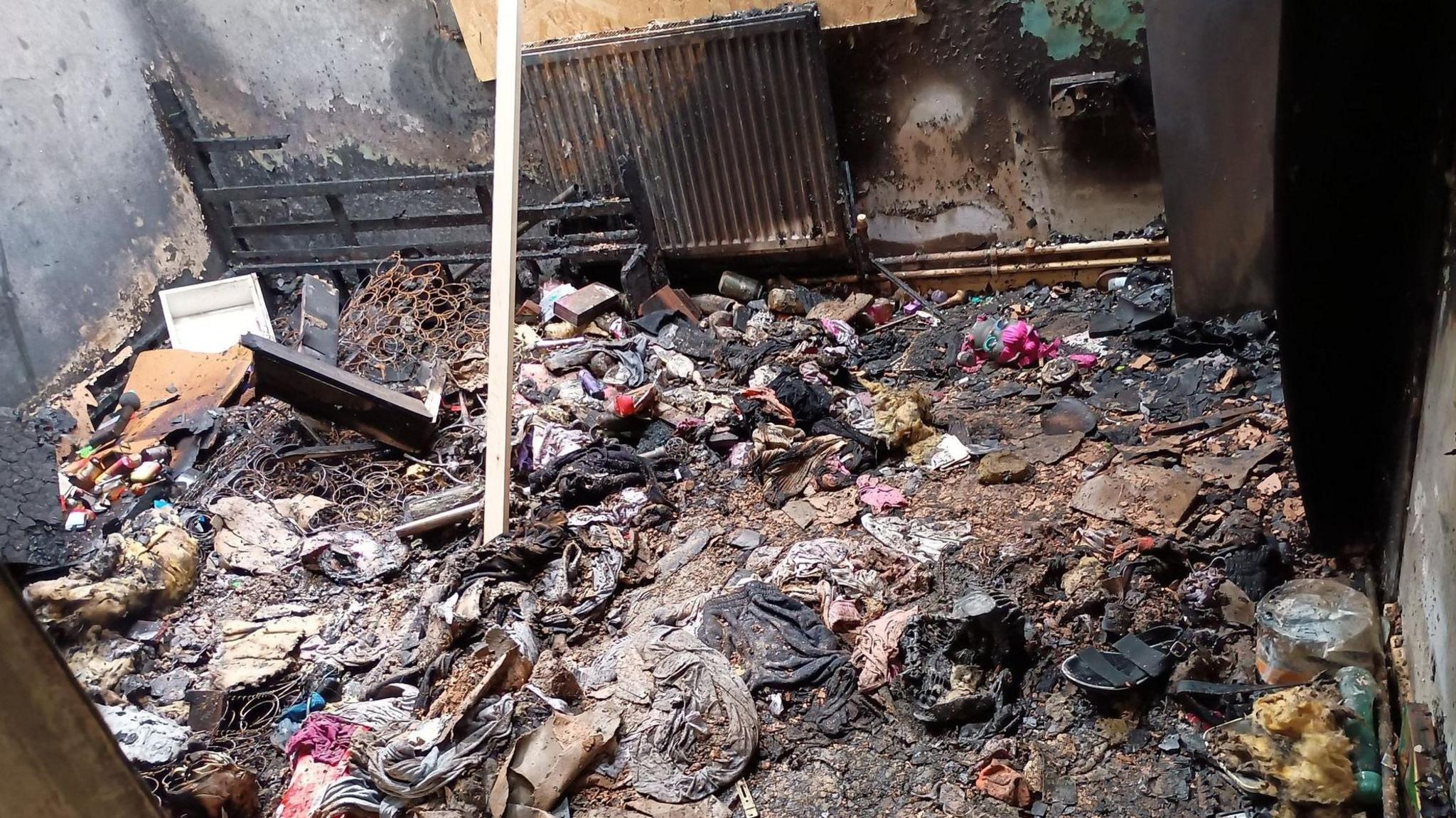 A family home after a fire with debris strewn around the floor including clothing, shoes and the springs from a burnt mattress. The walls and radiator are blackened from the flames.