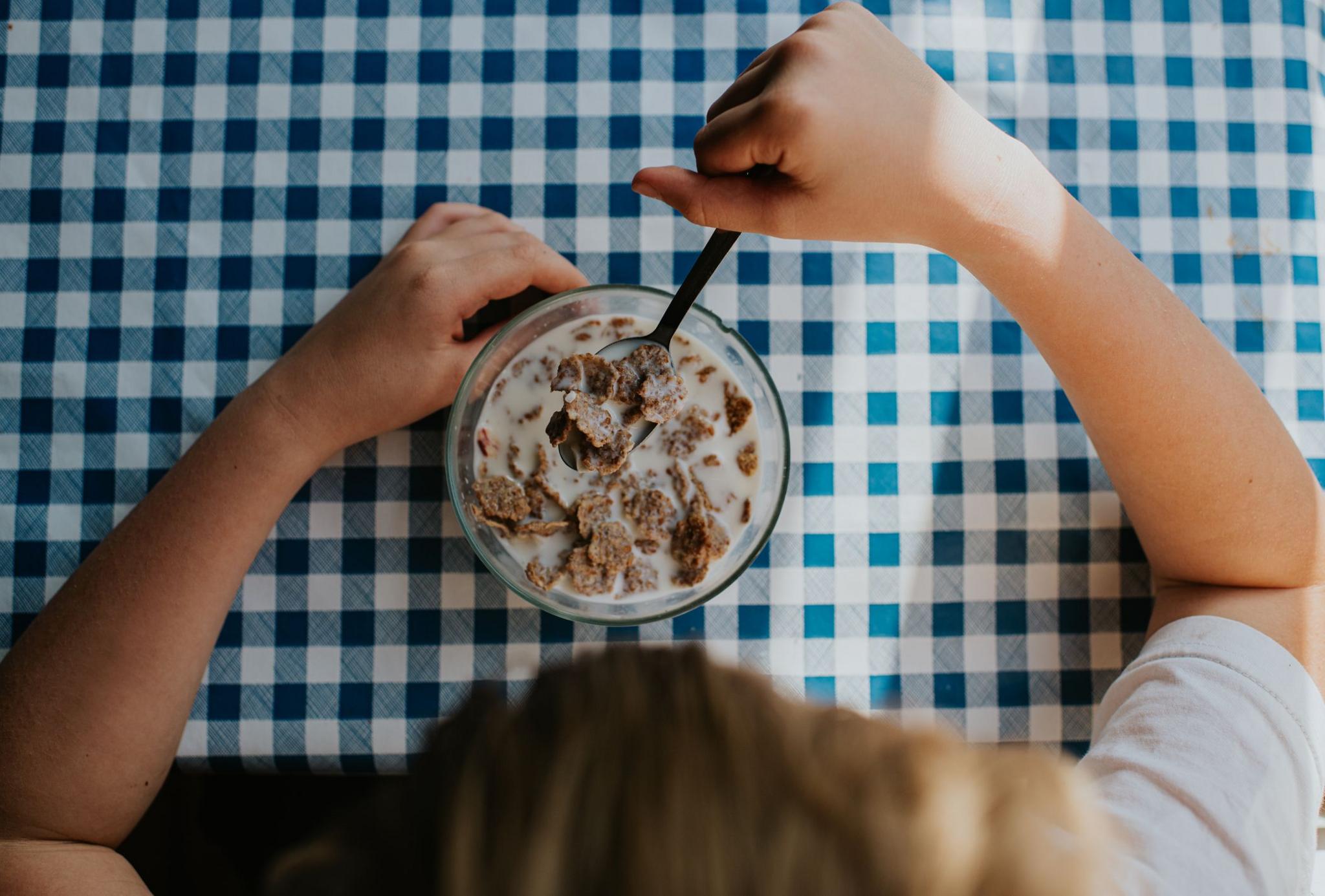 a child eating cereal and milk out of a bowl with a spoon, the bowl is on a chequered table cloth