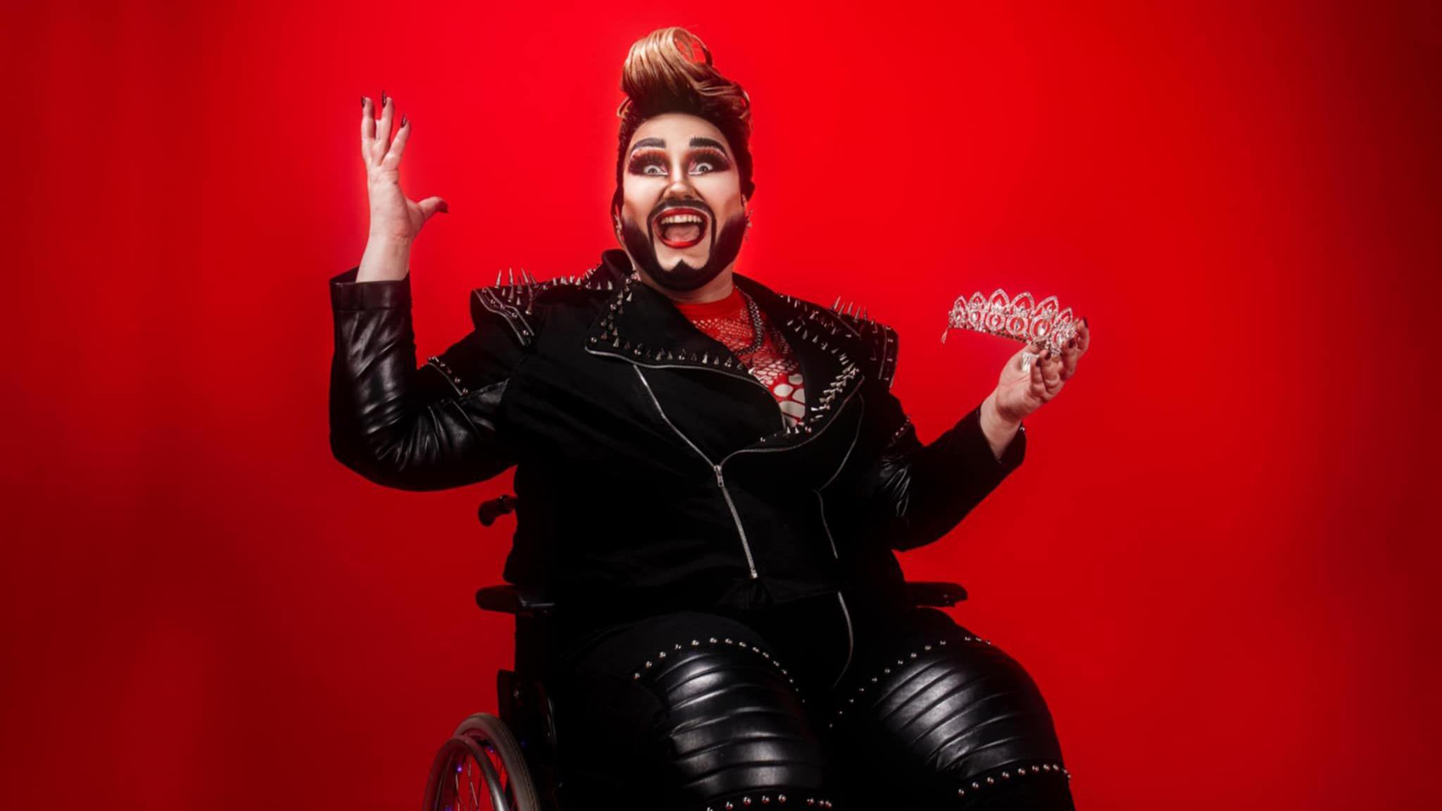 Drag artist Emma Round sitting in a wheelchair while wearing a black leather biker jacket and trousers. They are holding a tiara and sitting in front of a red backdrop.