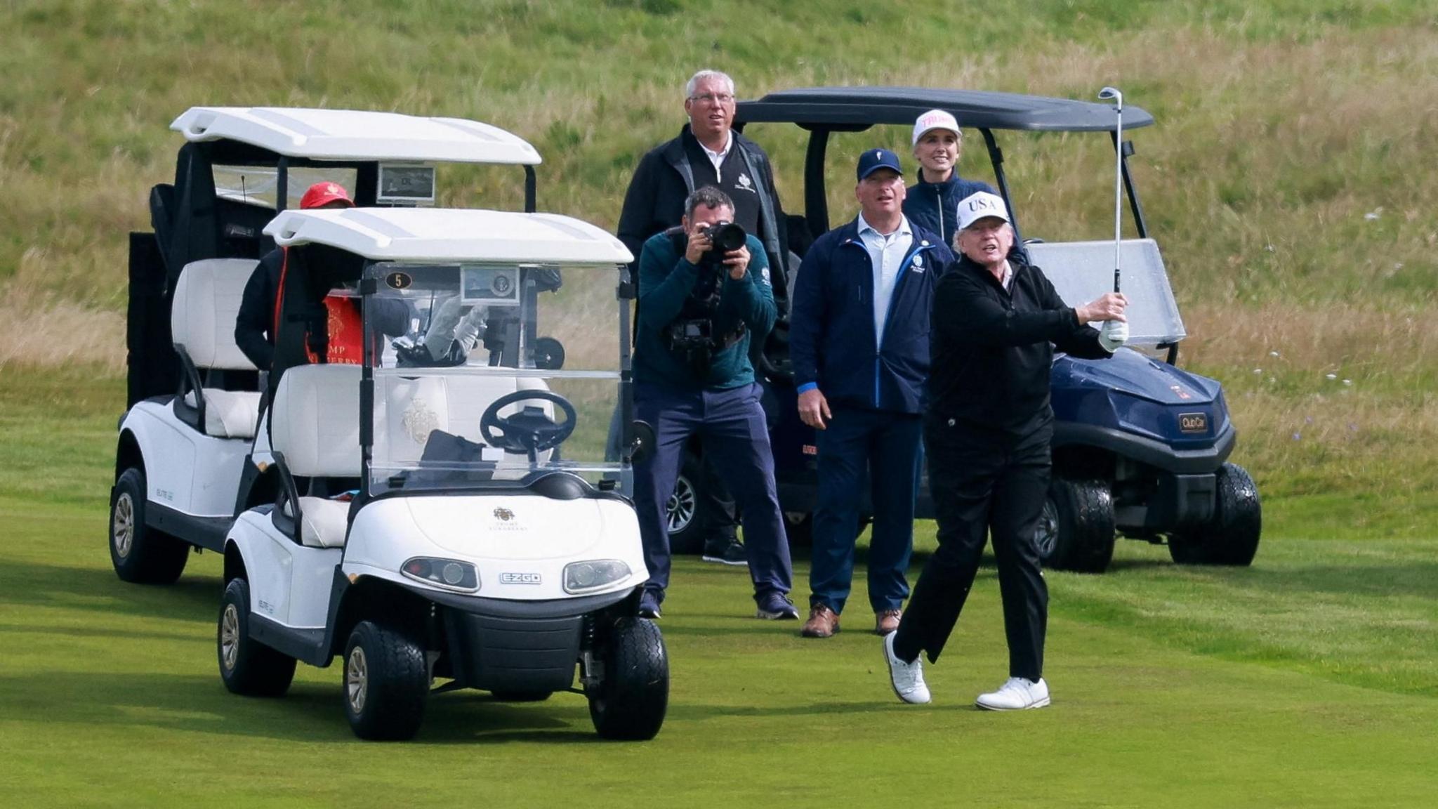 President Donald Trump at the end of his golf swing. He is standing next to three golf buggies and there are four peopel behind him watching, including one photographer.