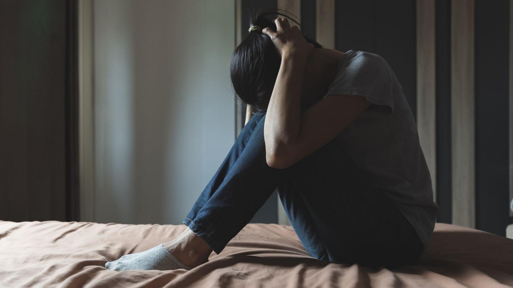 A woman with dark hair, a grey t-shirt and jeans sits on a bed with her head in her hands