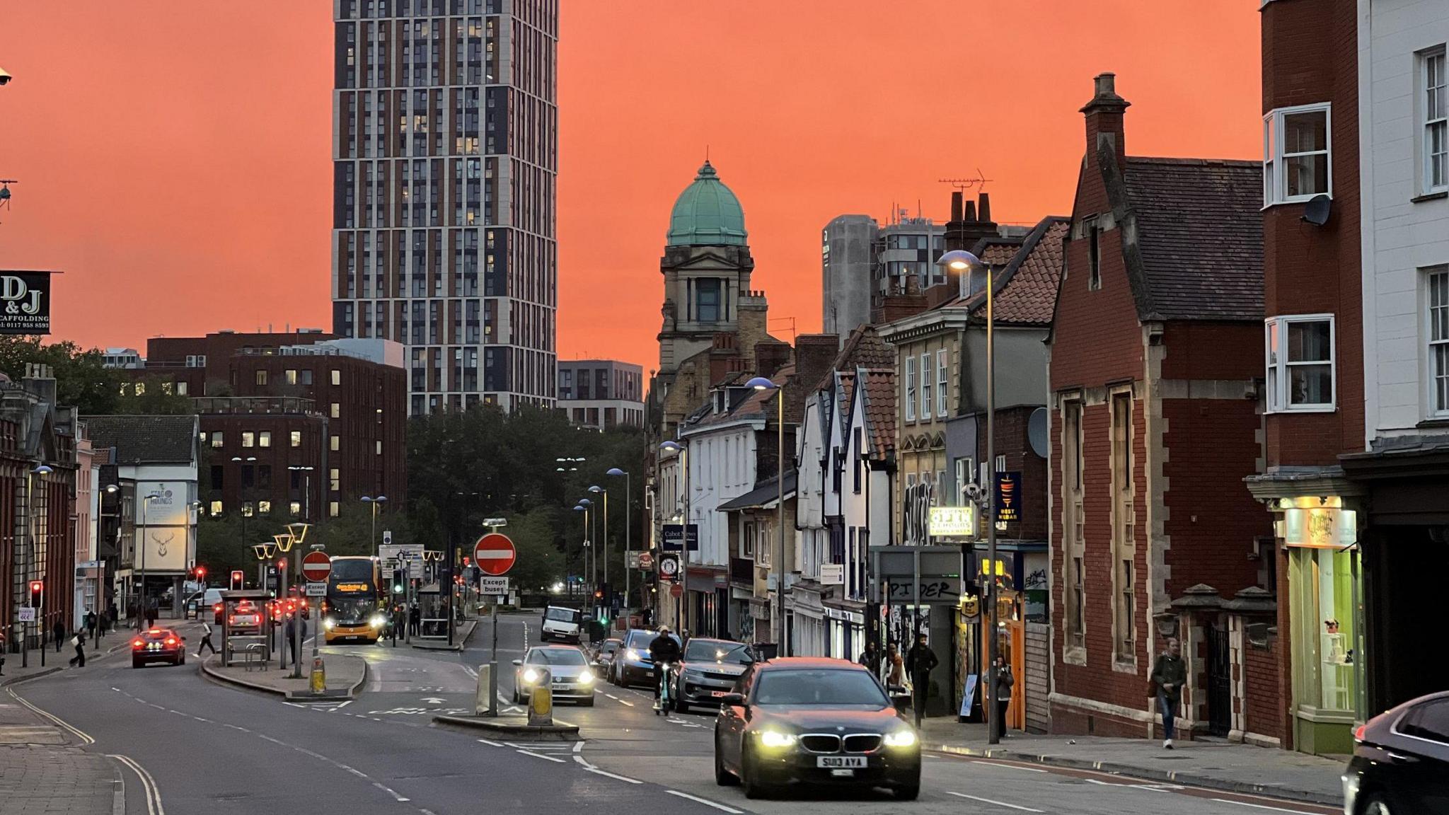 Old Market in Bristol is seen at sunset. The sky is an orange hue and cars are driving towards the camera with their headlights on, and street lights are also illuminated in the background.