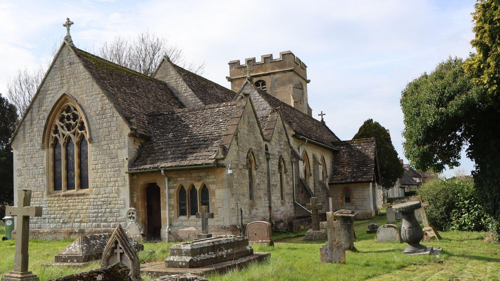 A view of a traditional English church on a sunny, with gravestones surrounding it. It is built in a light stone with Cotswold stone tiles
