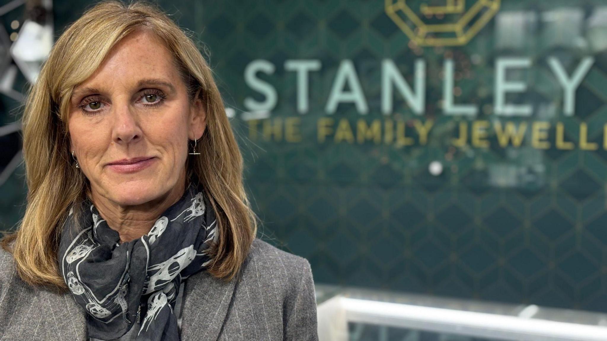 A woman is wearing a black and white scarf and grey blazer is looking into the camera. On a green-patterned wall behind her are the words "Stanley" and "The Family Jewellery". 