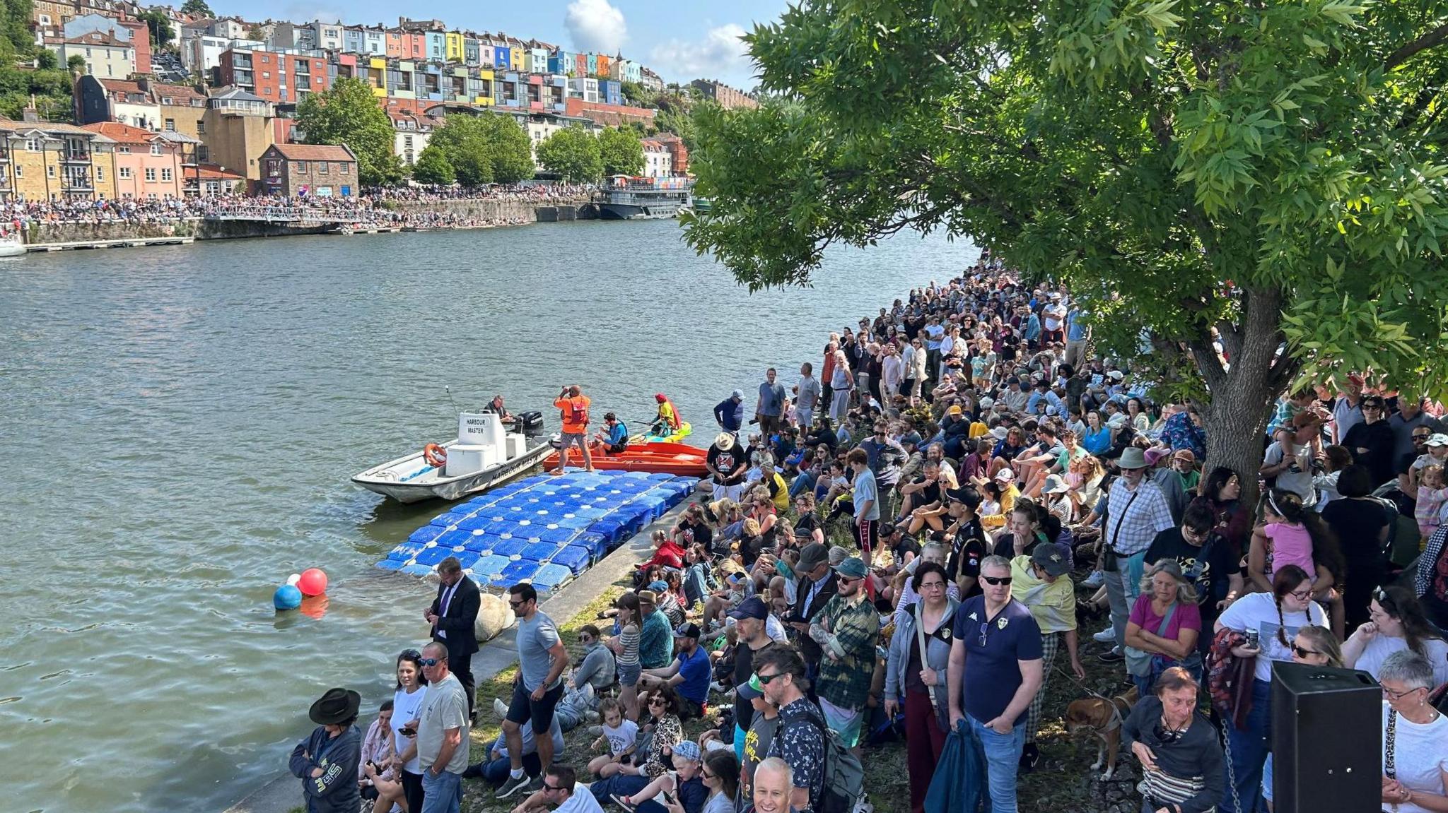 Large crowds watch the Bristol Birdman competition from the side of Bristol Harbour. In the distance are colourful houses of Cliftonwood