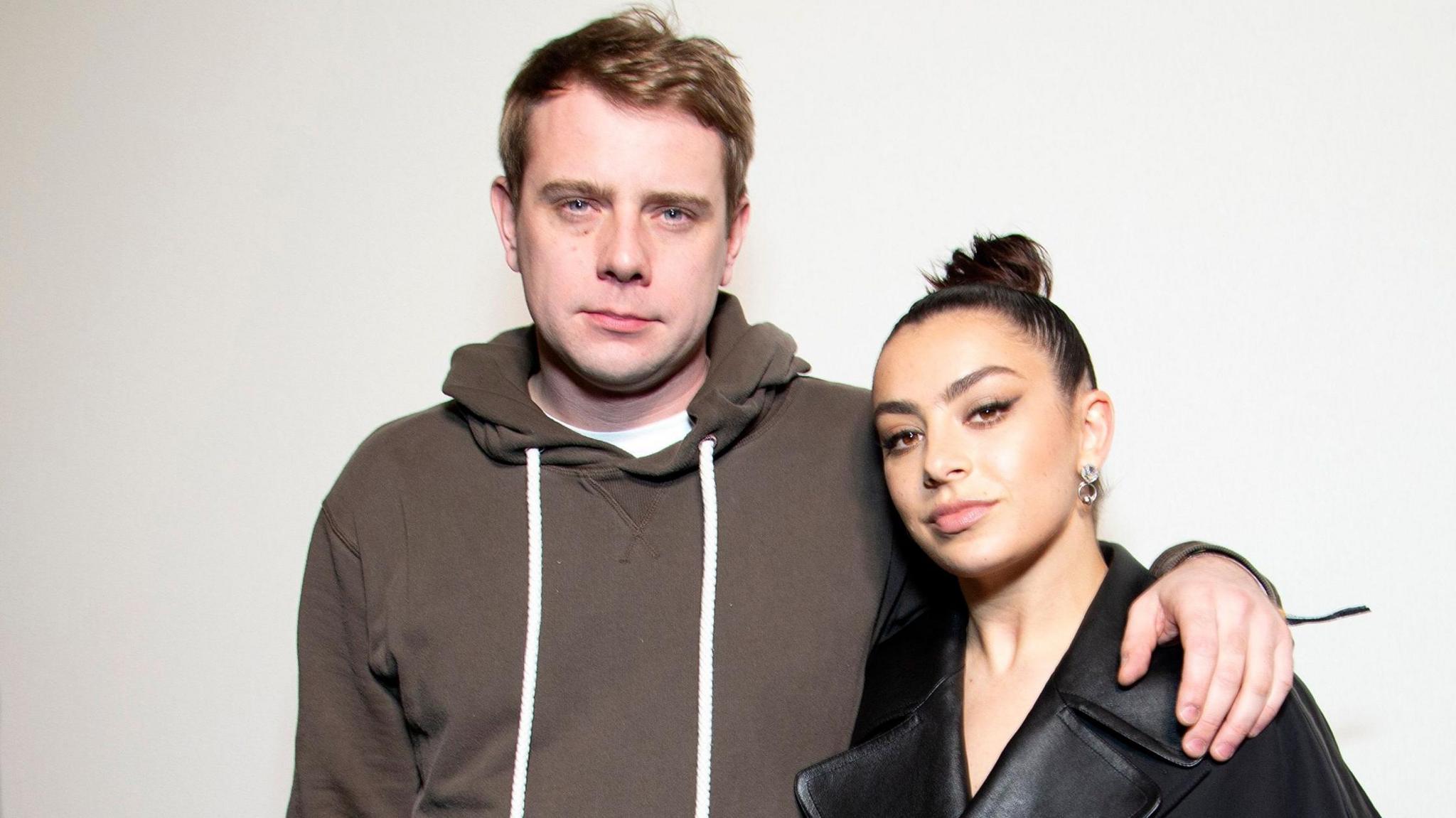A man with light brown wearing a brown hoodie with white drawstrings and a white T-shirt underneath, looks into the camera. He has his left arm around a woman wearing her dark hair in a tight bun. She is wearing a dark top and large earrings.