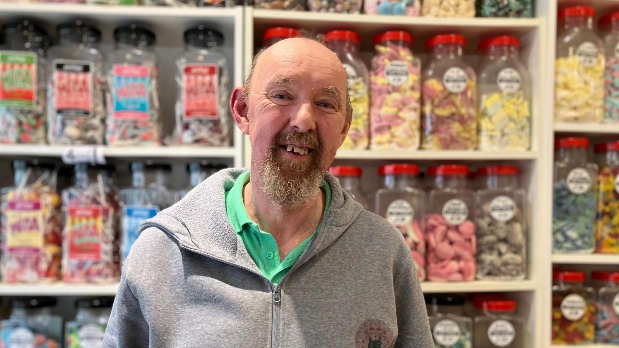 Steve Barnes is wearing a grey zipped hoodie and stands in front of shelves holding many different jars of brightly coloured sweets.