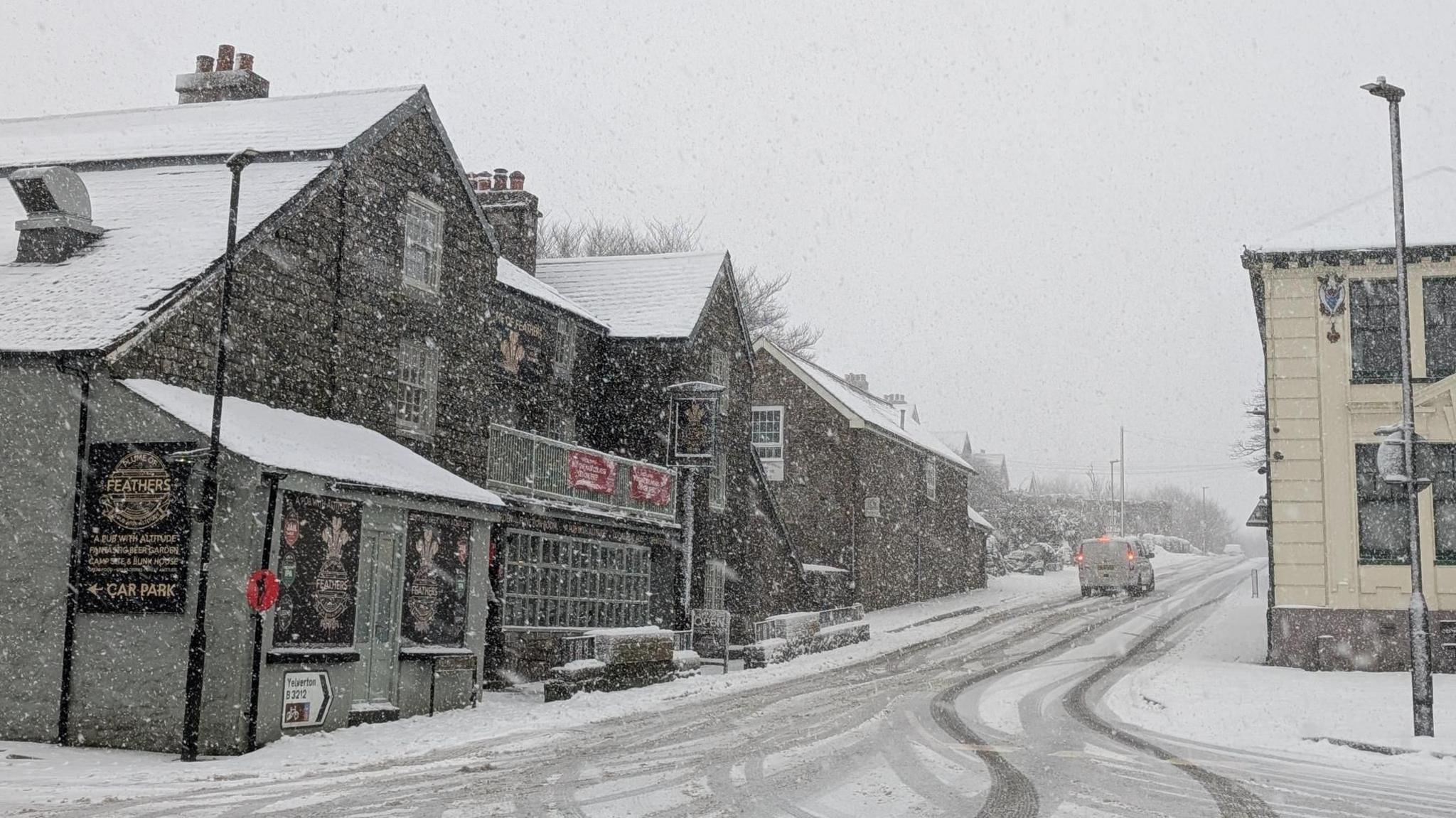 Snow is seen on the road with car tyre marks. There is a cream coloured building on the right side and other buildings on the left. The sky is grey.