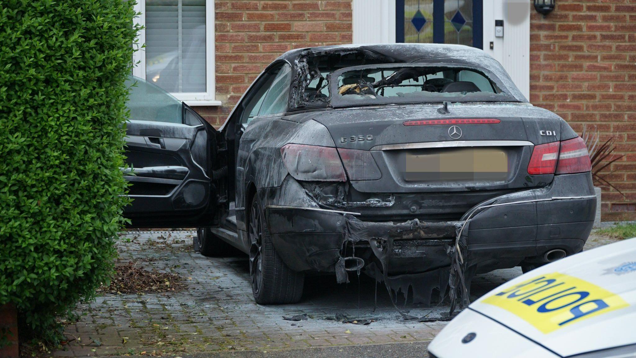 A black burnt out Mercedes sports car sits on the damaged driveway of a semi-detached house in Eastbourne with a police car parked in front of it