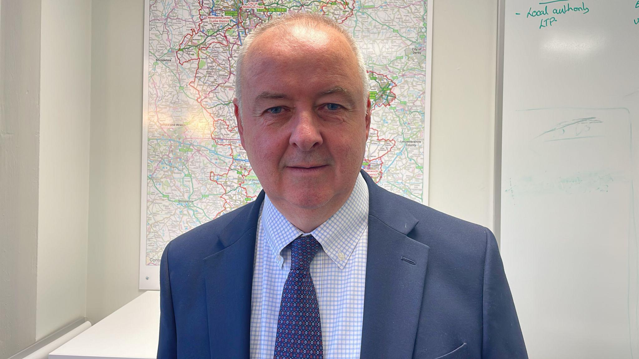 A man with very short grey hair is wearing a dark blue suit jacket with a dark blue patterned tie, and white shirt with light blue squares on it. He stands in front of a whiteboard and a map of Staffordshire