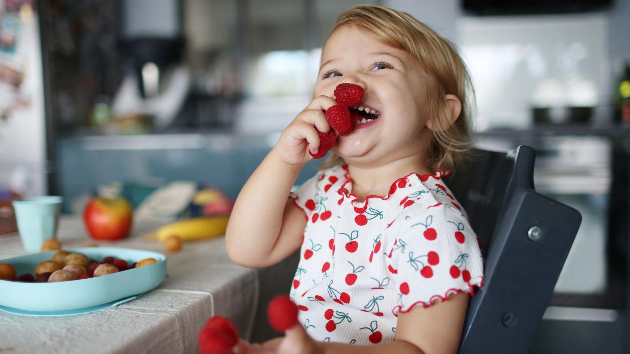 A child with blonde hair smiling wearing a cherry printed top holding raspberries in both hands. 