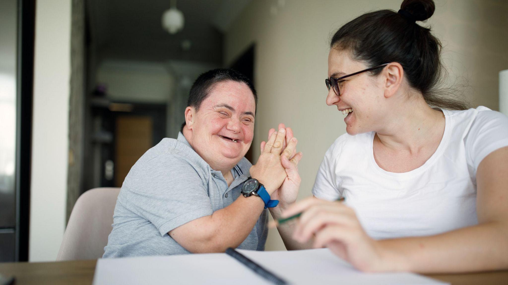 A man laughs with his female support worker while they are both sat at a desk with a pen and paper 