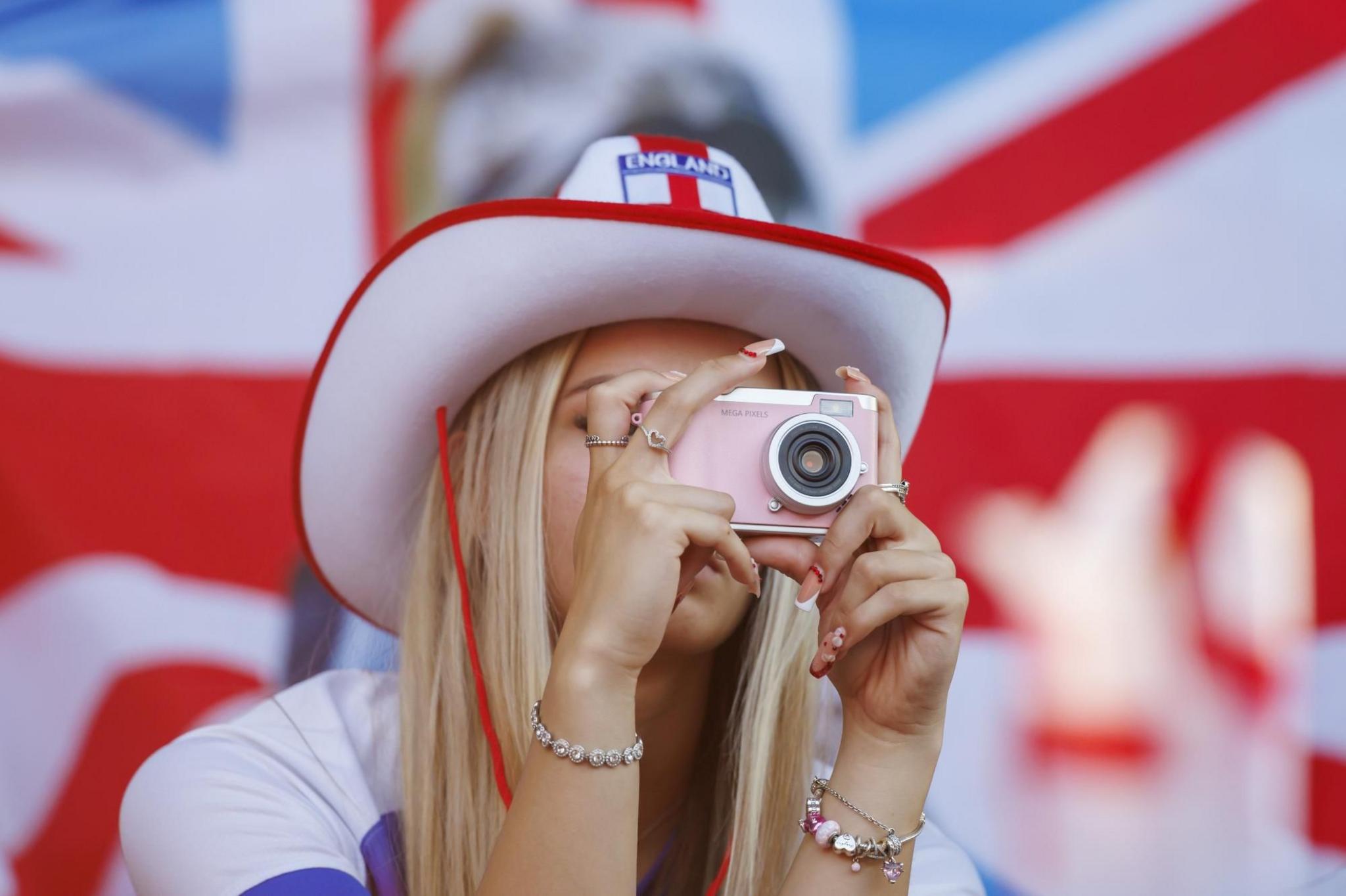 An England fan wearing a cowboy hat in the colours of the English flag takes a picture with a pink digital camera. She has long nails with sparkling red beads.