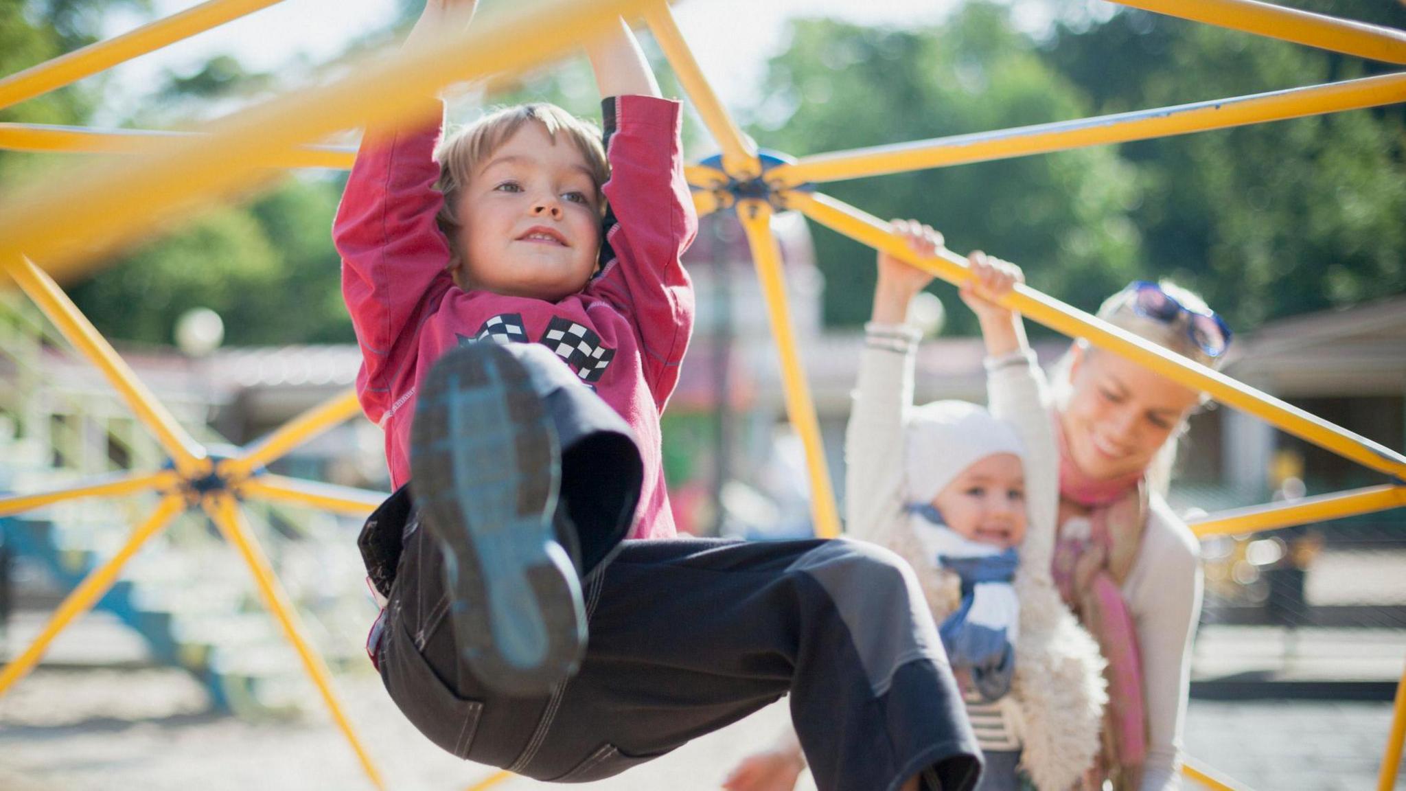 A young boy wearing blue jeans and a red sweatshirt swinging from a climbing frame. In the background a woman is holding another young child who is hanging from the climbing frame.