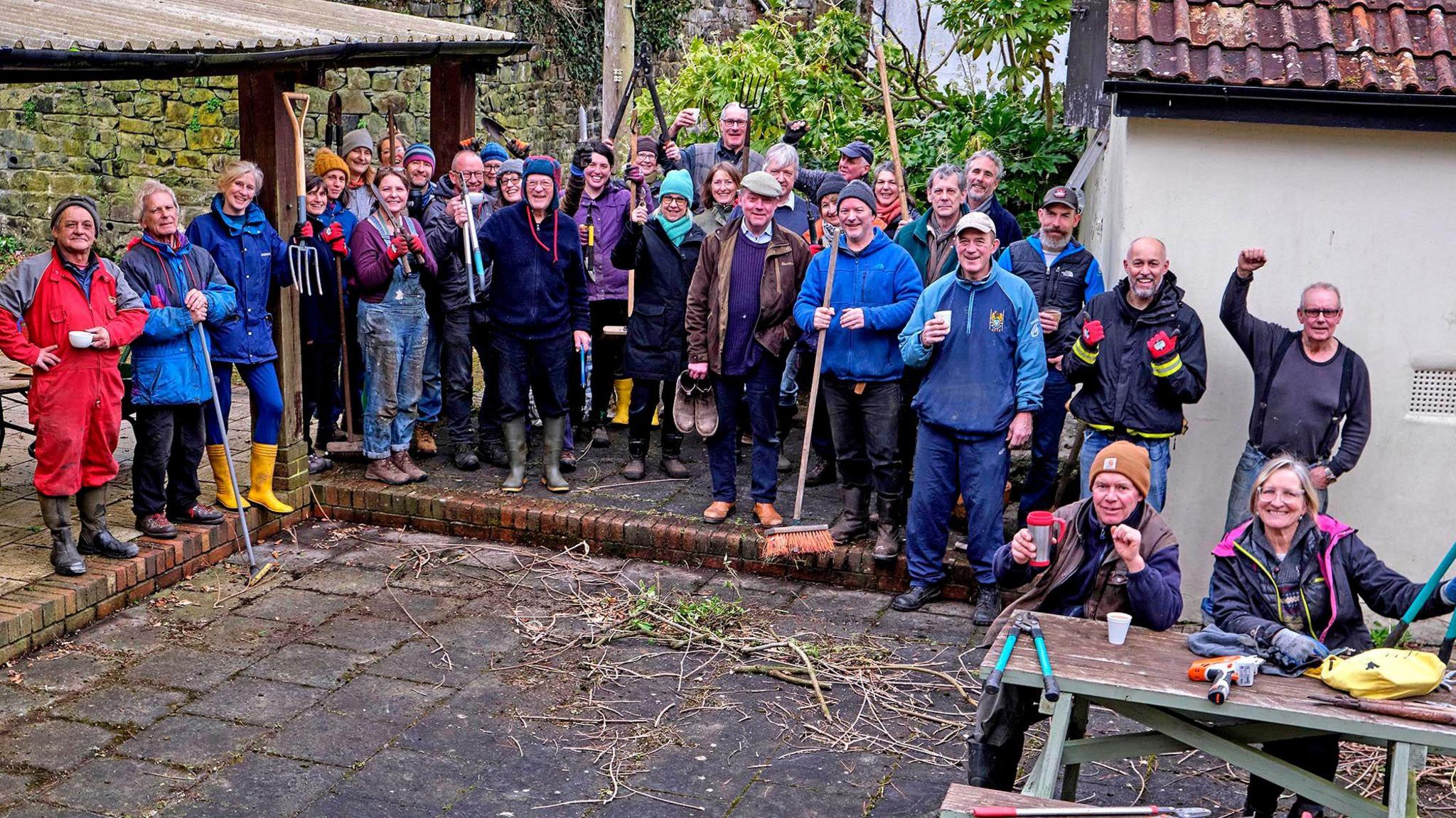 The Drewe Arms community stand outside the pub. The group, made up of more than 30 people, are standing and looking toward the camera. They are dressed in hats and coats. They are holding outdoor equipment including brooms and pitchforks. 