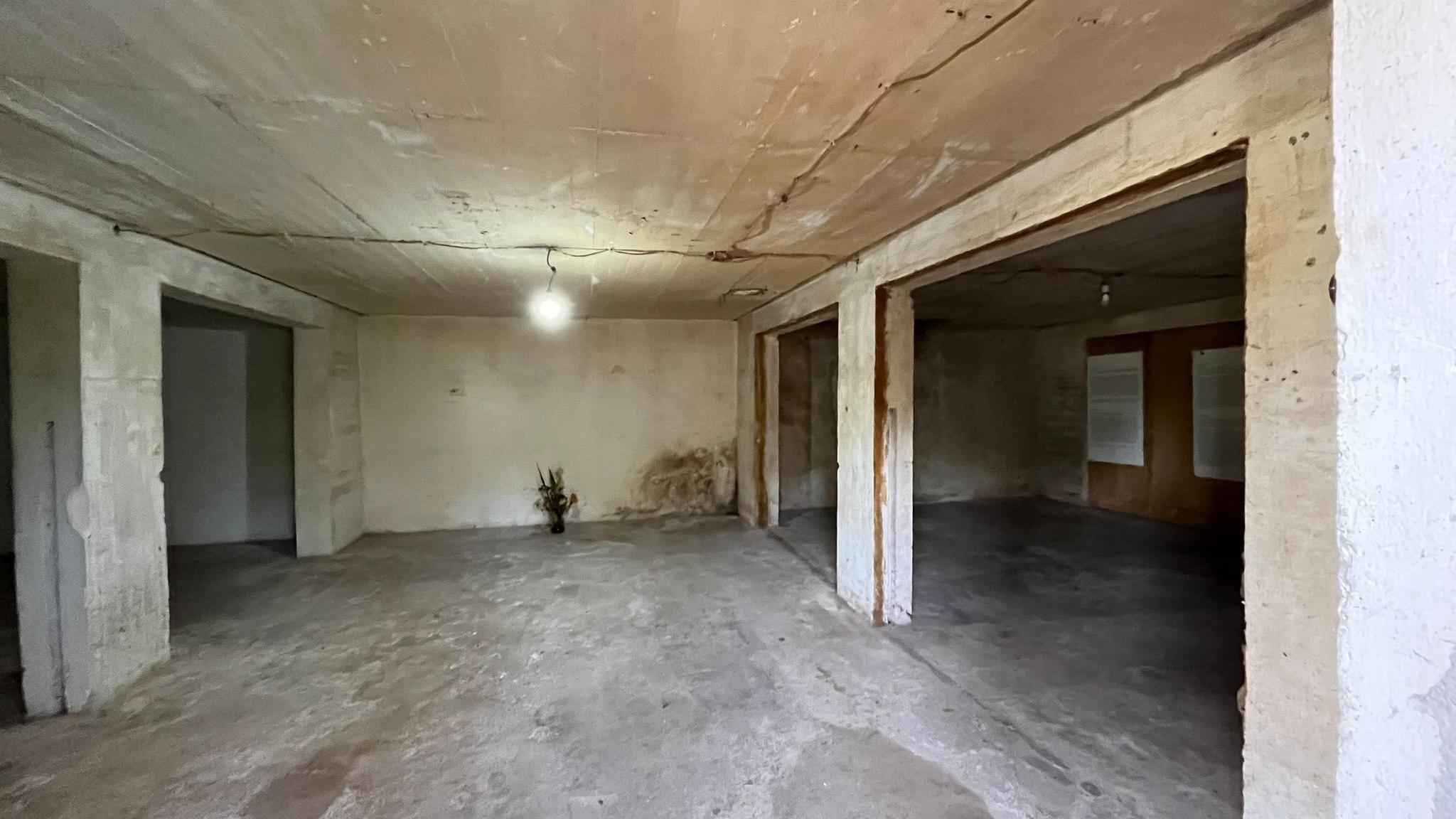 The inside of the potato warehouse: the floor is concrete and a single lightbulb hangs from the ceiling. Concrete pillars hold up the ceiling and what appears to be a dry bunch of flowers stands inside a vase placed at the far end of the room 