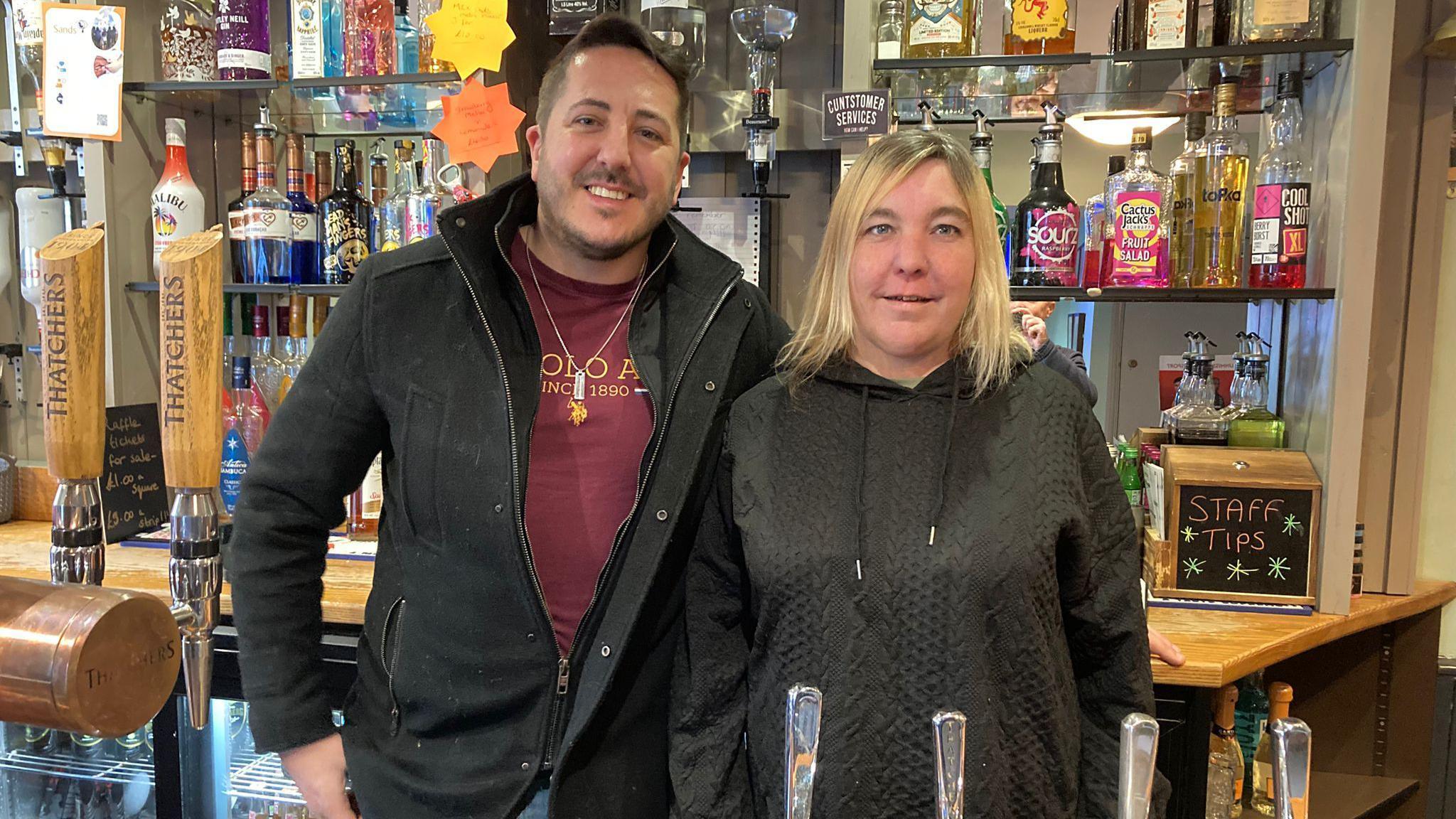 Dale Smith and manager Hayley Parsons stood behind the bar. Bottles of spirits align the back of the bar behind the pair and they are both looking into the camera smiling.