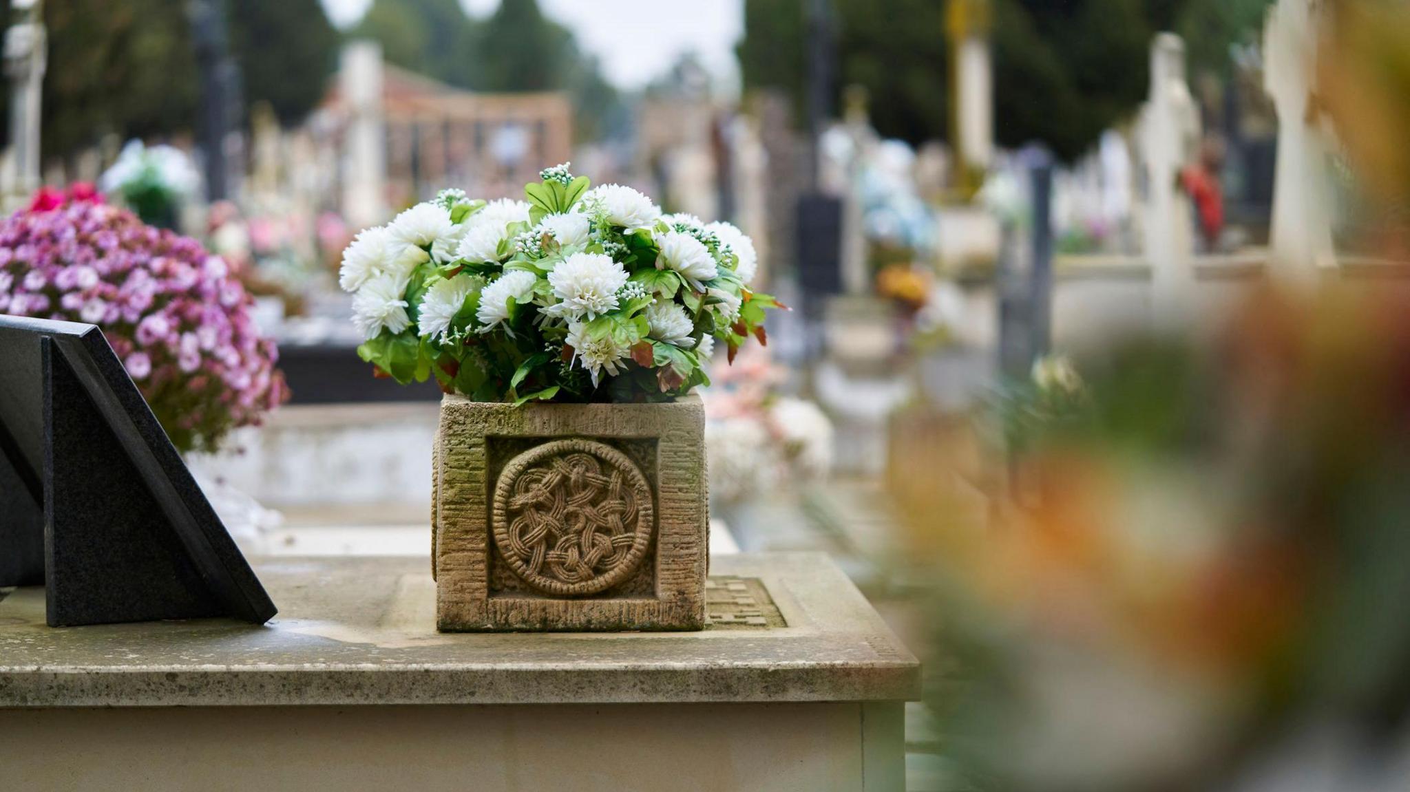 The picture shows a basket of flowers in a cemetery seen from a side angle to the graves in a row with several flowers in the background.