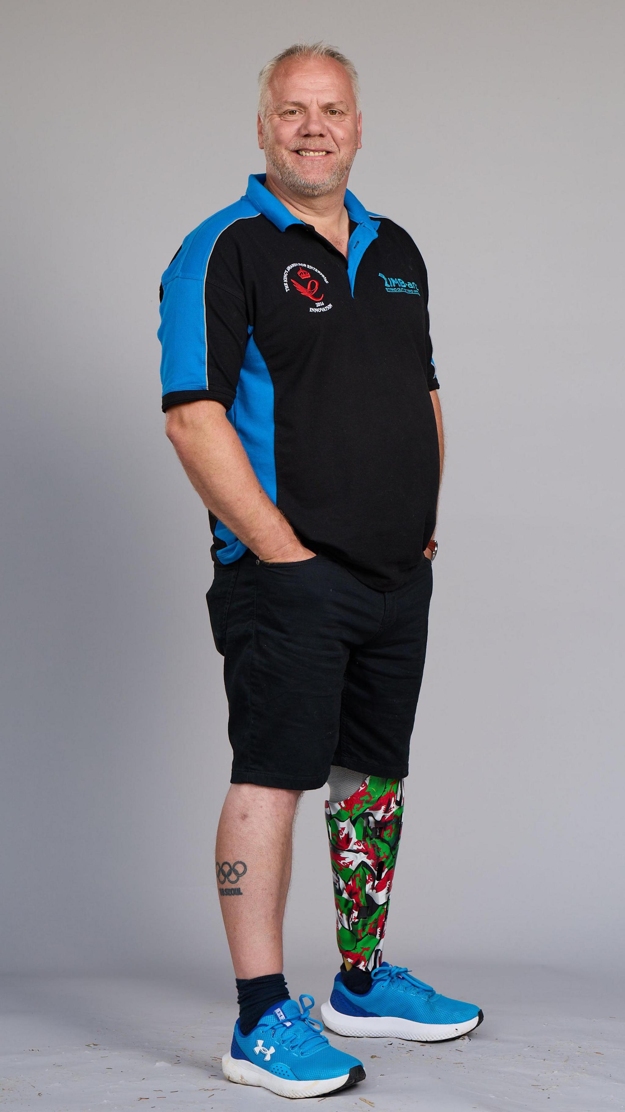 Mark poses for a full length photo in a studio. He has close-cropped grey hair and a stubbly beard. He's wearing a black and blue T-shirt with his company logo, black shorts and blue trainers. He has the Olympic rings tattooed on his right leg, and his left prosthetic leg has a cover attached to it mimicking the shape of a lower leg. The cover is brightly patterned with the red, green and white colours of the Welsh flag.